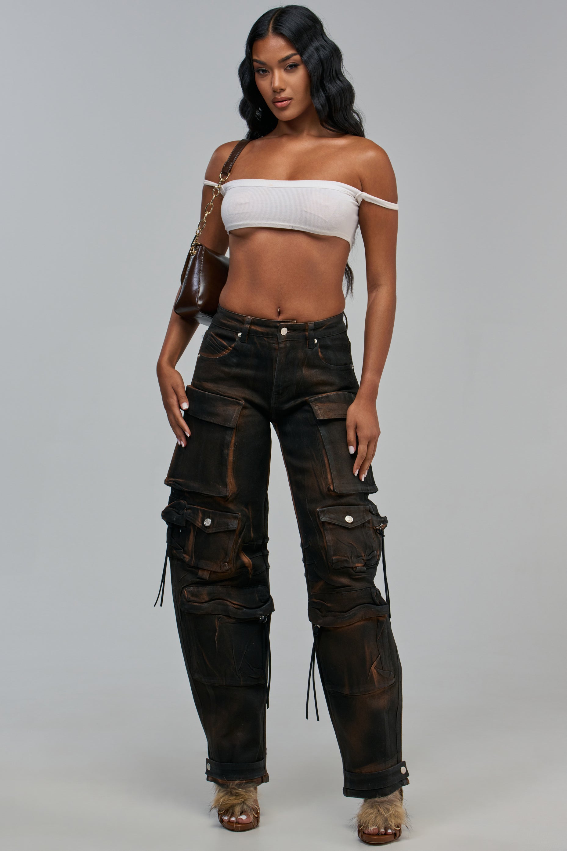 A person wears a white off-the-shoulder crop top, a shoulder bag, and DARK AND HEAVY CARGO PANT, which are dark, distressed brown denim pants with a barrel leg silhouette.