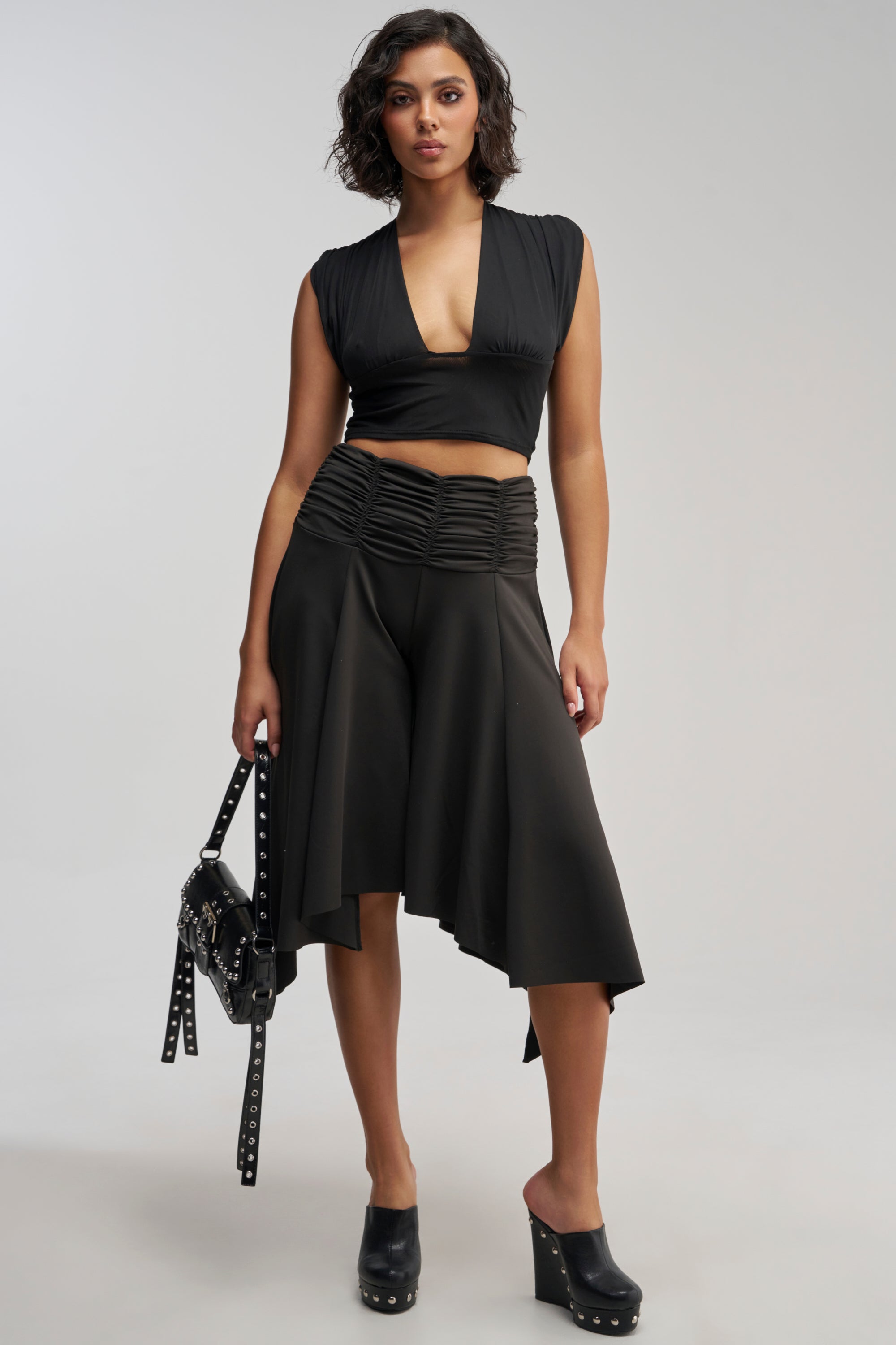 Woman in a black sleeveless crop top and asymmetrical skirt, holding a studded handbag and wearing black platform heels, standing against a plain light background. For a fresh, modern edge, her chic ensemble could effortlessly transition to include the FLYING AWAY GAUCHO pants.