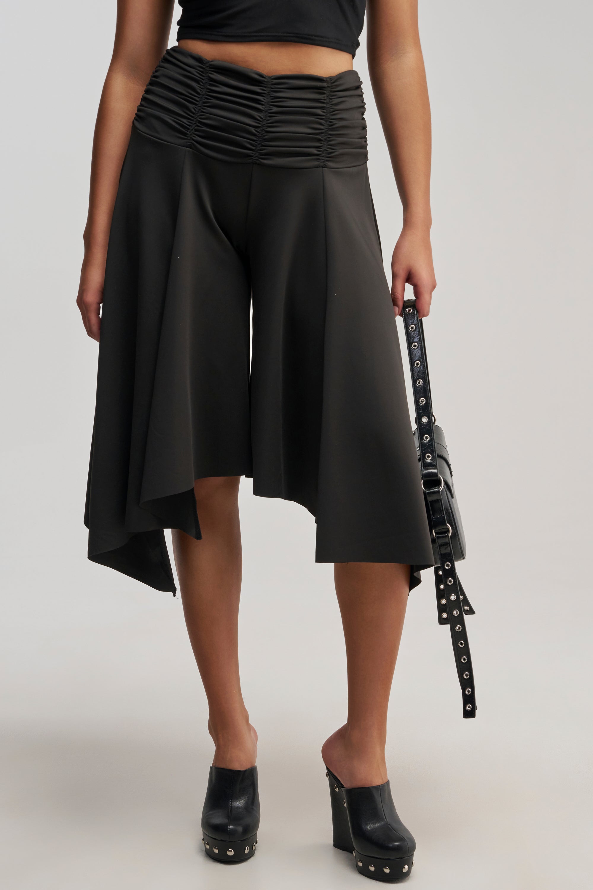 A person wearing the FLYING AWAY GAUCHO high-waisted, black, asymmetrical wide leg pants with ruching at the waistband, paired with black clogs and holding a black handbag with silver studs. Only the lower half of the body is visible.