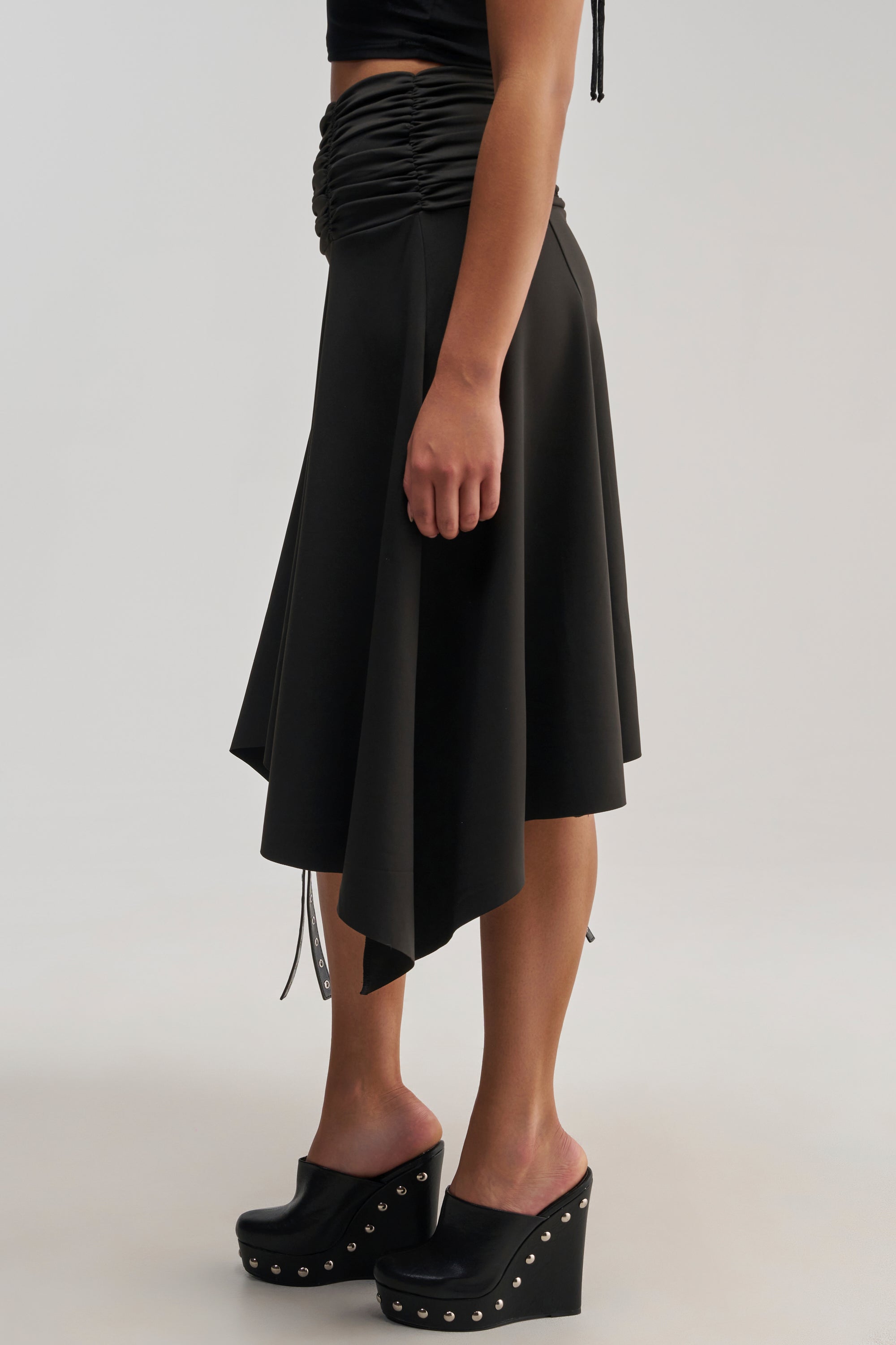 A person wearing the FLYING AWAY GAUCHO black wide leg pants styled in an asymmetrical midi cut, paired with a black ruched crop top and black platform clogs with studs, standing against a plain light background.