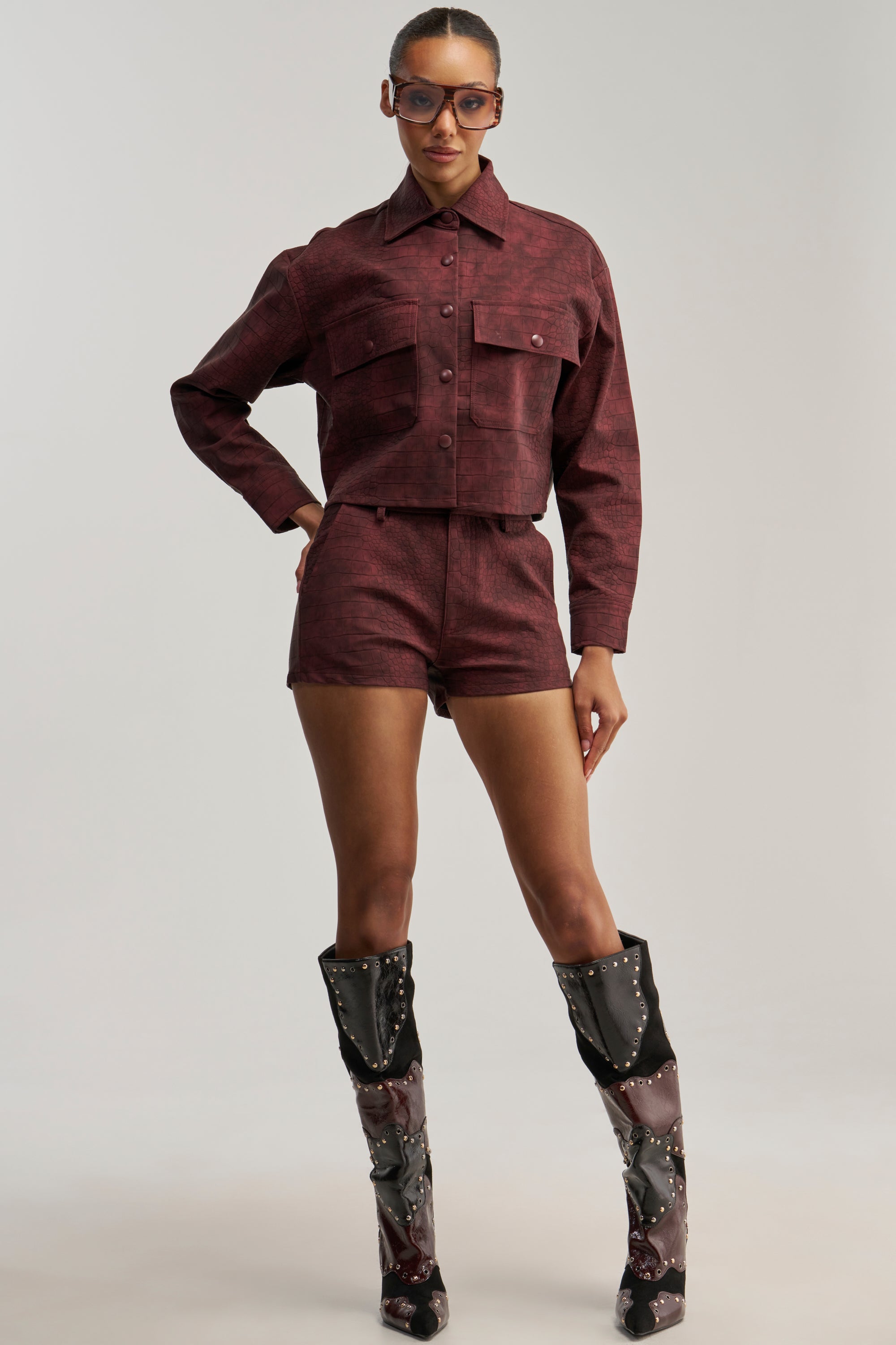 A woman stands confidently against a plain background, wearing the MOODY CROC SHORT—croc-embossed high-rise mini shorts—paired with a burgundy jacket and tall black and burgundy boots adorned with silver studs.