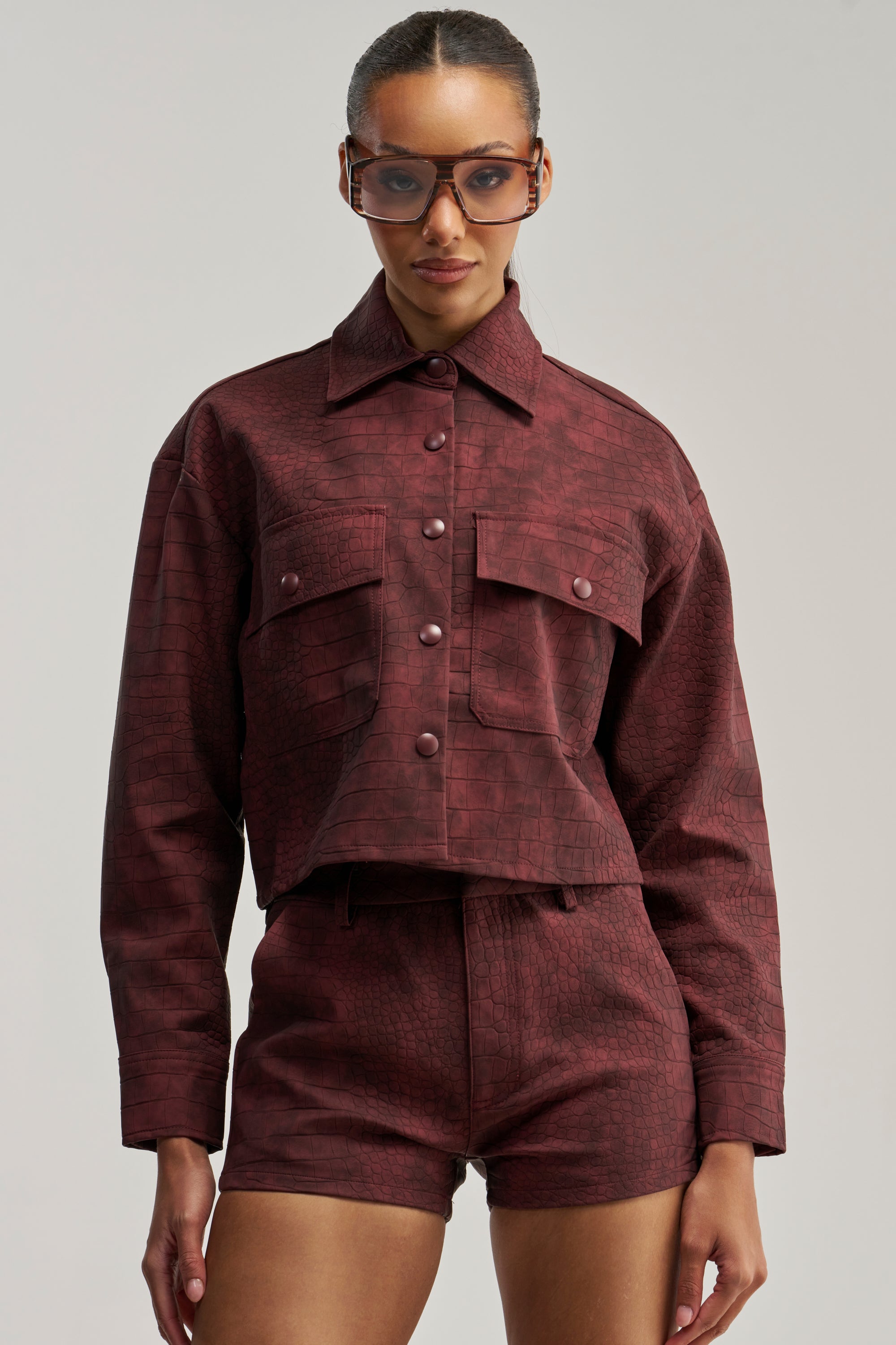 A woman wears the MOODY CROC LONG SLEEVE BLOUSE, a textured burgundy crocodile-patterned button-down jacket with large buttons, paired with matching high-waisted shorts. Her hair is slicked back and she accessorizes with stylish sunglasses, set against a plain light-colored background.