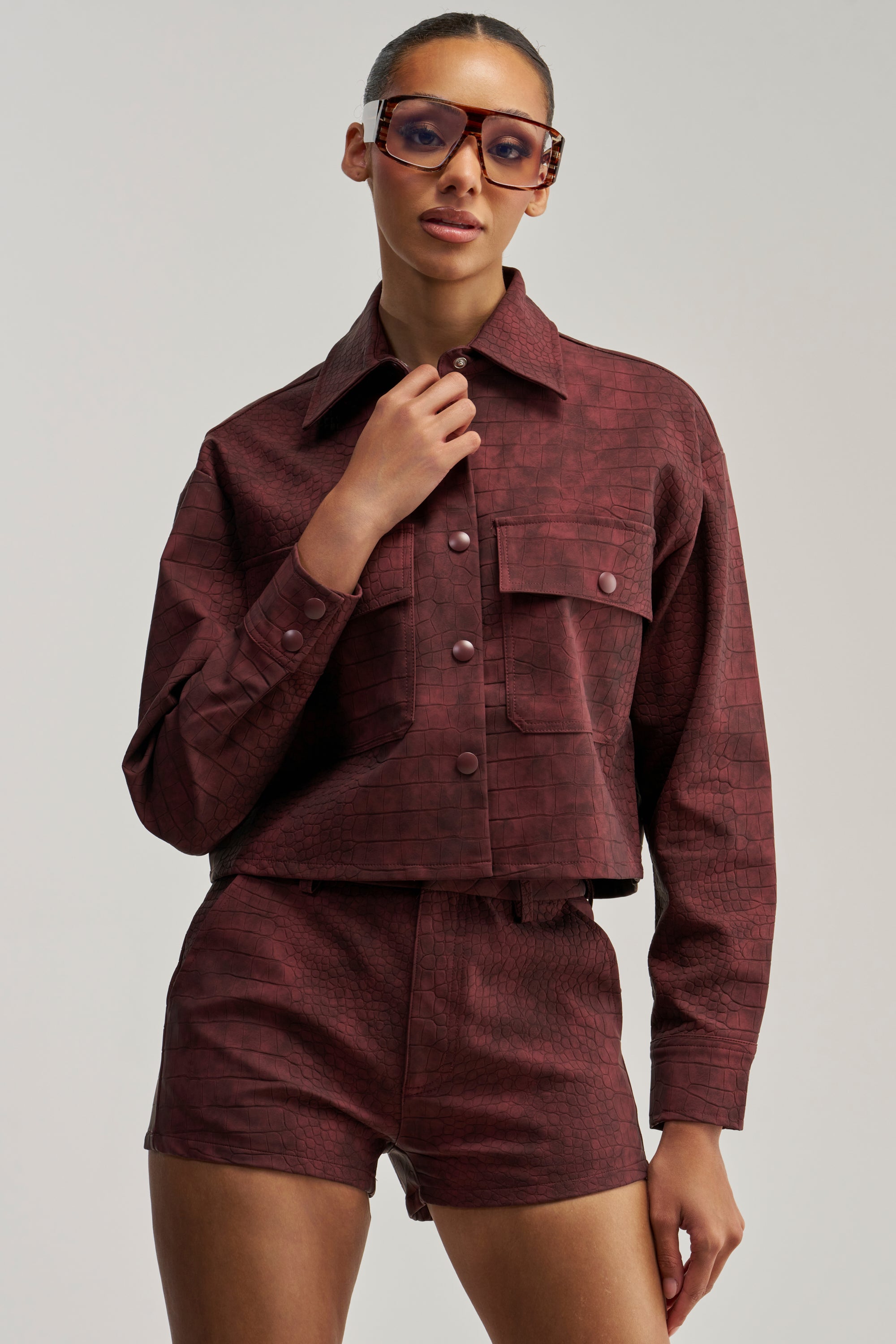Woman wearing large sunglasses and the MOODY CROC LONG SLEEVE BLOUSE, styled as a textured maroon button-up jacket with matching high-waisted shorts, poses against a plain light background.