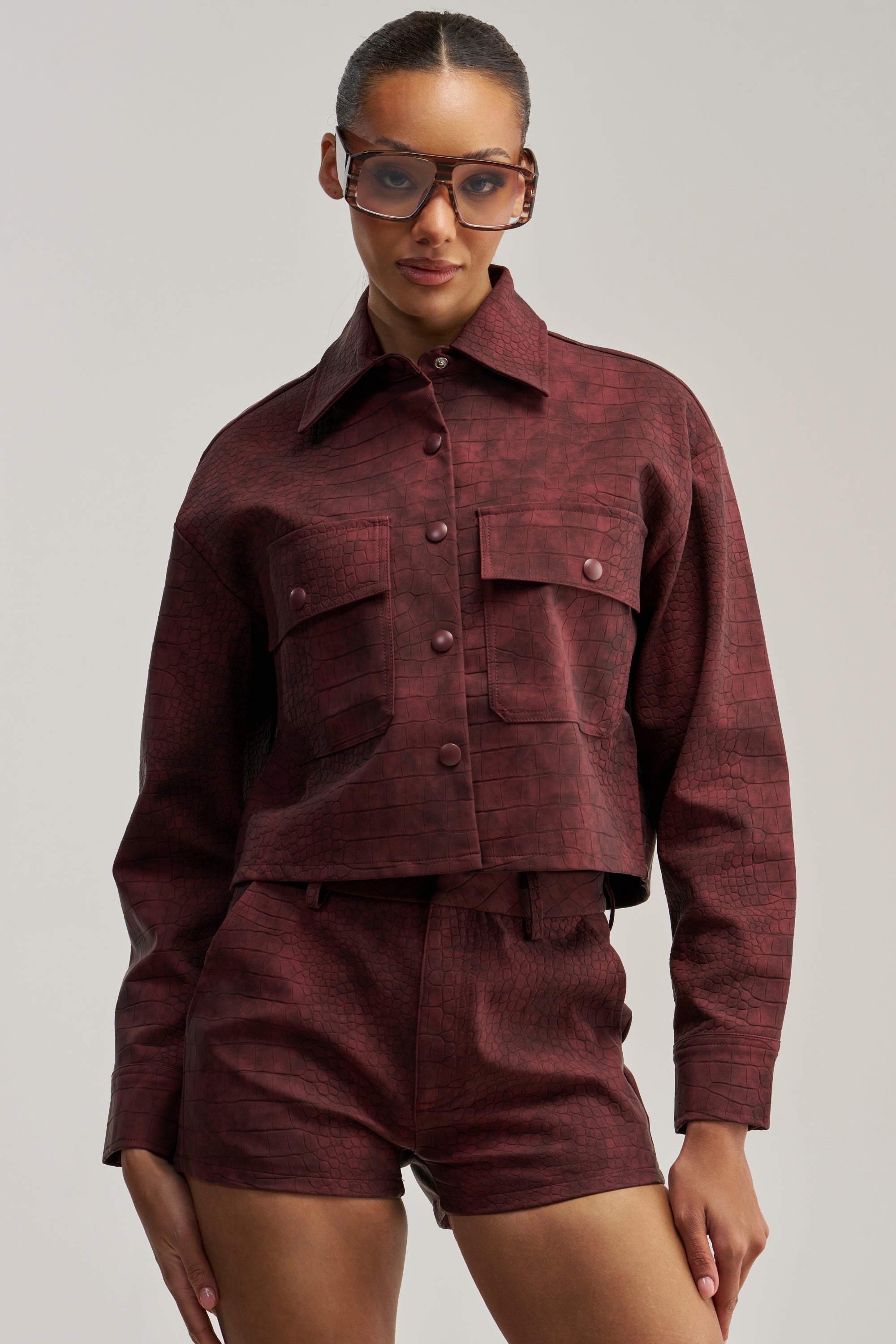 A woman wearing the MOODY CROC LONG SLEEVE BLOUSE with a matching maroon, textured jacket and shorts set, styled with oversized sunglasses and large front pockets, stands against a plain background.