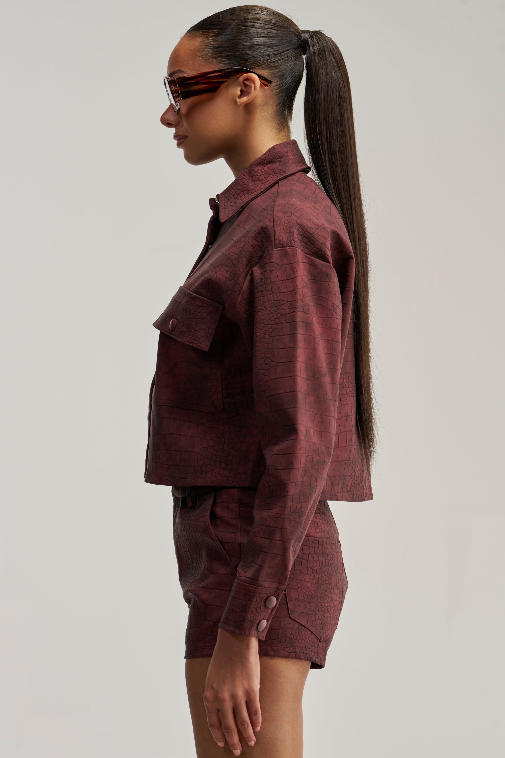 Woman in a maroon textured jacket and matching shorts, standing in profile with a long ponytail and sunglasses against a plain background—a stylish look elevated by her MOODY CROC LONG SLEEVE BLOUSE layered beneath the jacket.