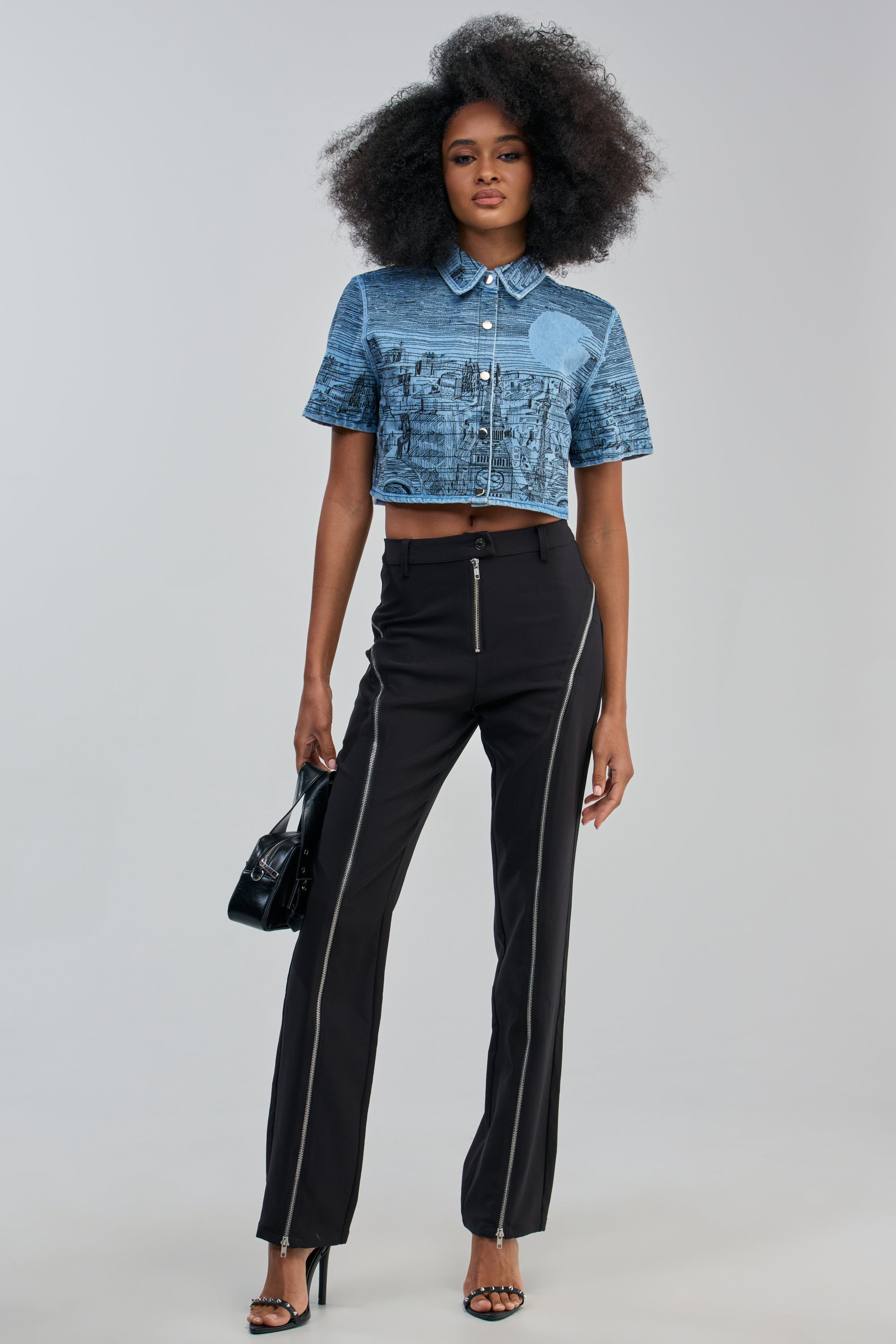 A woman with voluminous curly hair stands against a plain background, styled with edgy-chic flair in a blue cropped shirt and the EASY LOVER TROUSER—a black sculpted zipper trouser featuring a high-rise fit—paired with heeled sandals and a small black handbag.