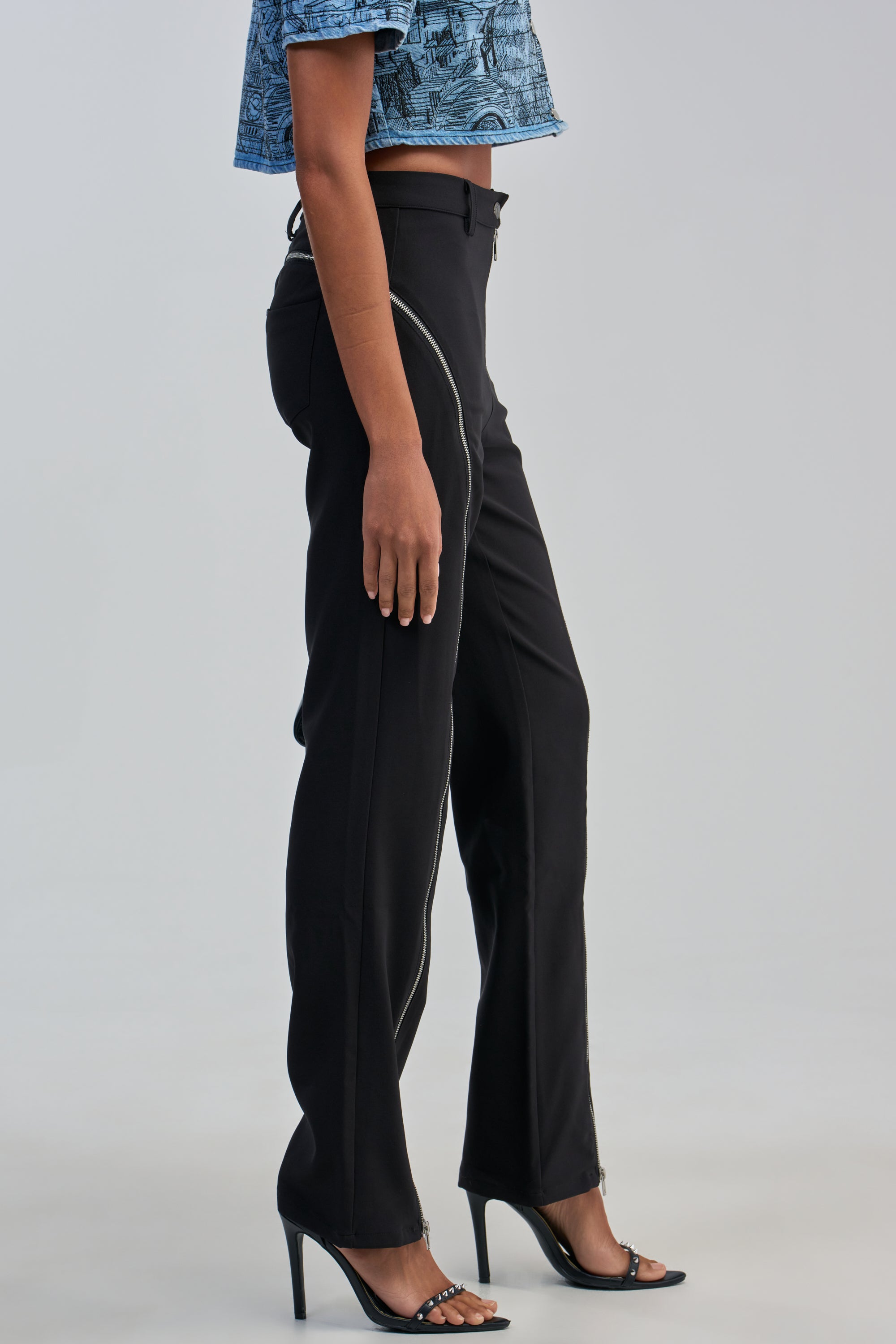 A person wearing the EASY LOVER TROUSER, featuring a sculpted zipper design and high-rise fit, paired with black high-heeled sandals and a blue patterned cropped top, stands against a plain light background for an edgy-chic styling.