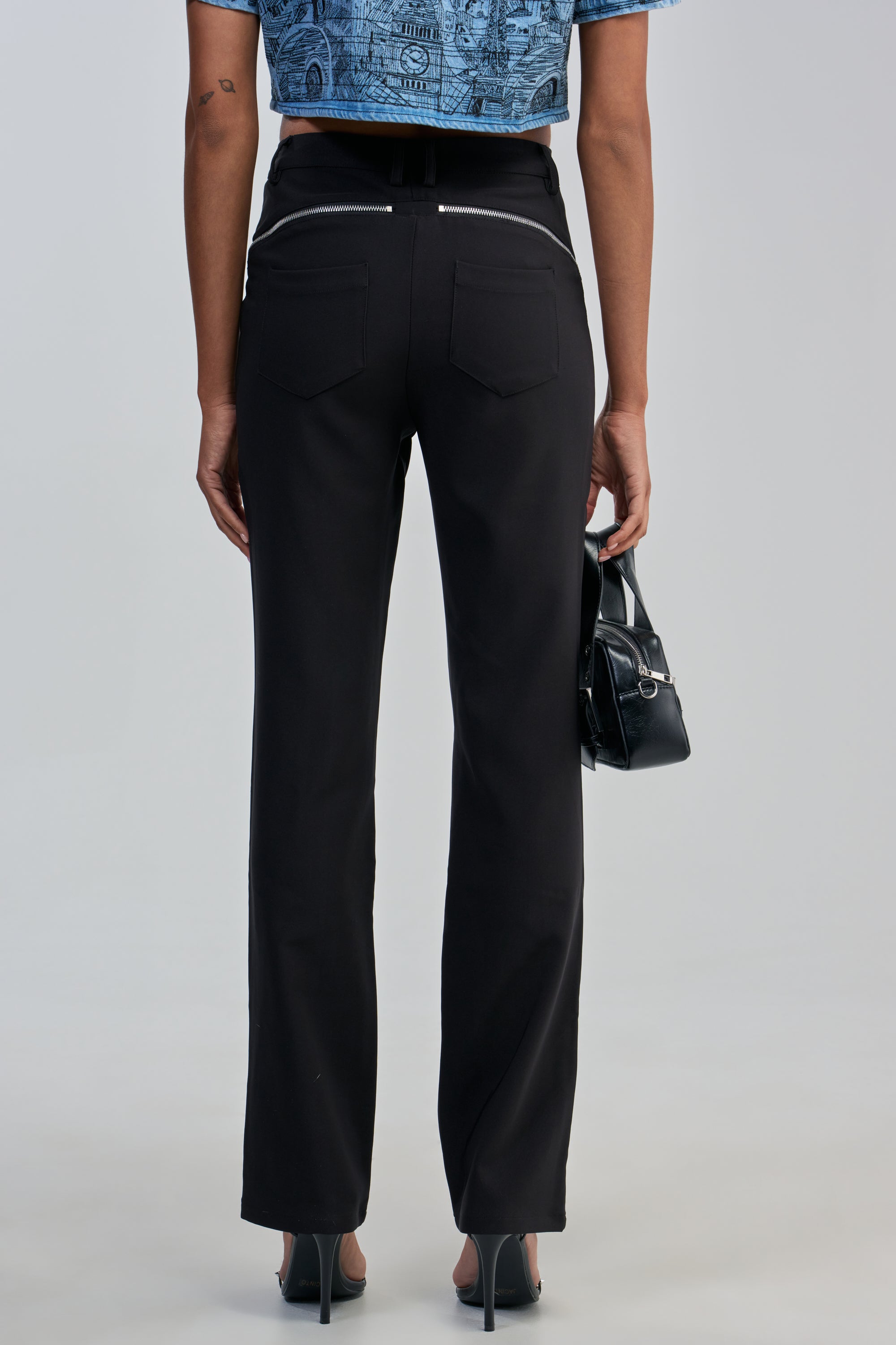 Dressed in the EASY LOVER TROUSER, this person showcases edgy-chic style with black high-waisted pants, a blue patterned crop top, and black heels. Posed from the back while carrying a small black handbag against a plain light-colored backdrop.