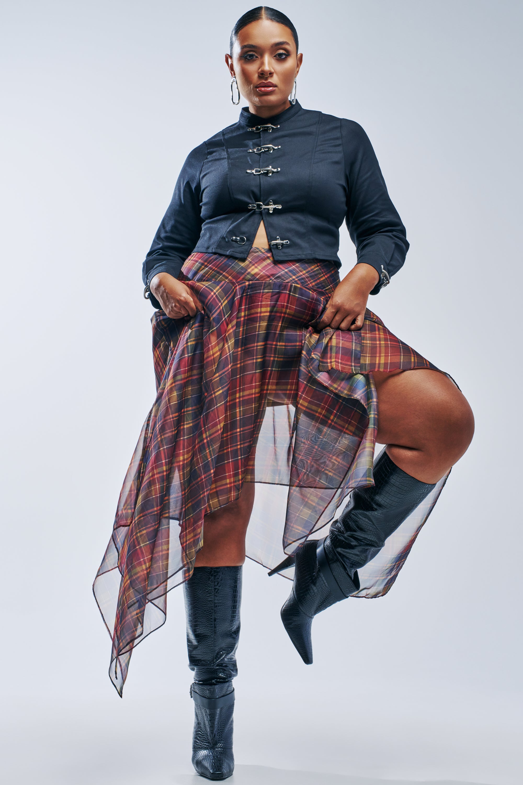 A person wearing a cropped black jacket with silver buckles and a MIXED FEELINGS PLAID SKIRT, styled with a sheer, pleated chiffon handkerchief hem and black knee-high boots.