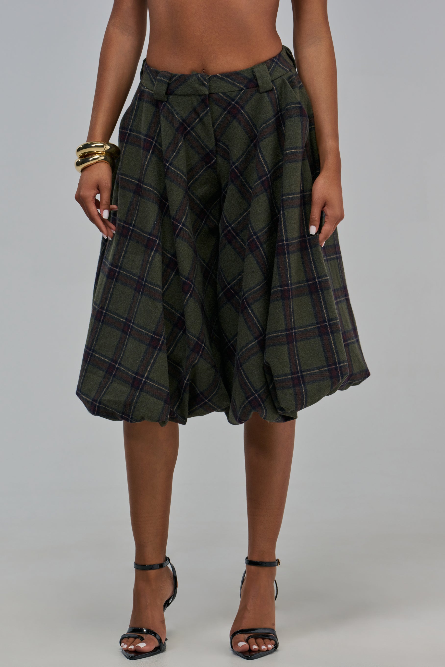 A person wearing COOLEST ON THE BLOCK PLAID BERMUDA SHORT high-waisted, olive green plaid gaucho pants with a pleated, voluminous silhouette and black ankle-strap heels.
