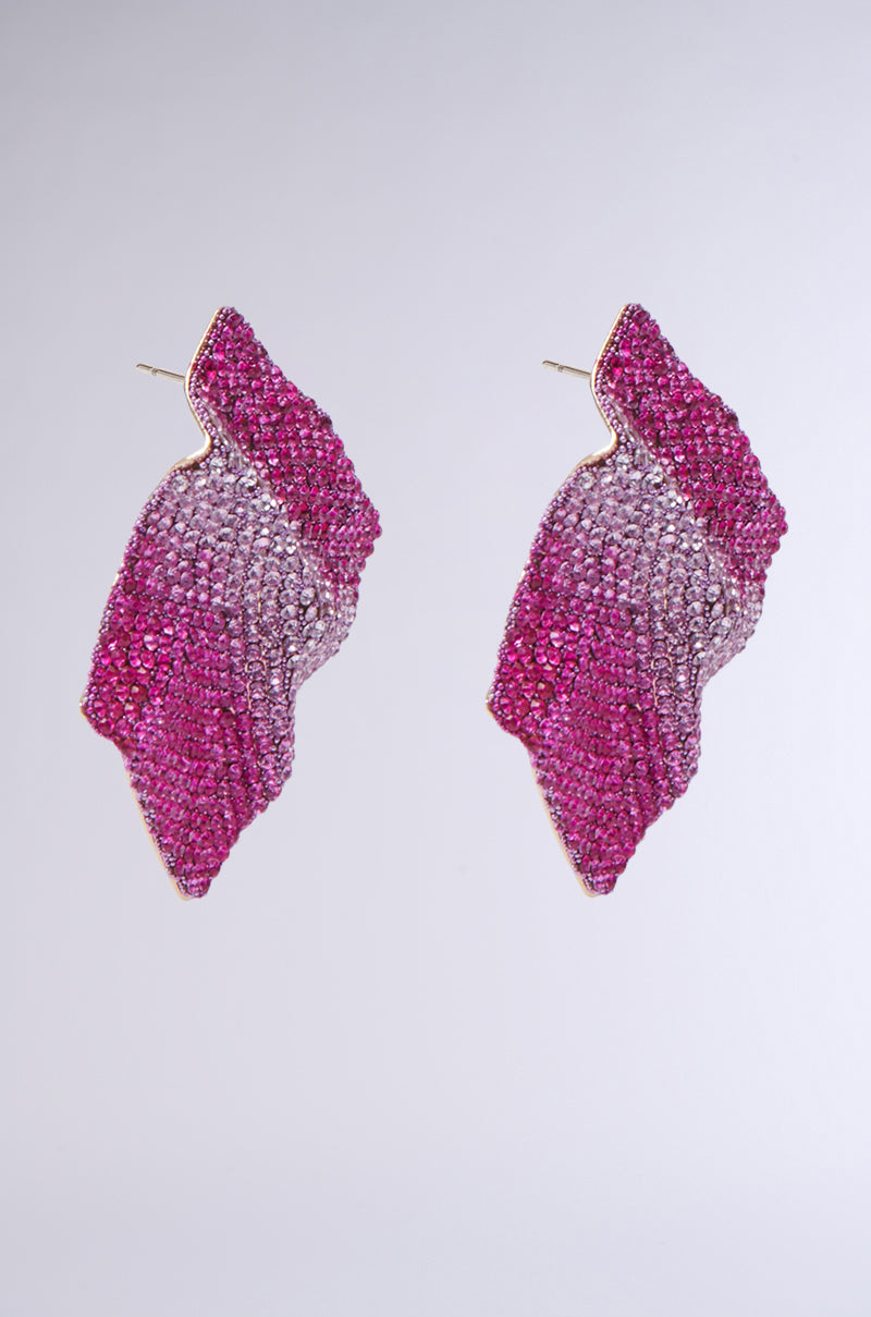 The ITS MY WORLD BLING EARRING features a pair of shimmering, leaf-shaped earrings adorned with small pink and silver crystals that create a gradient effect. These earrings showcase a unique wavy design outlined in gold, with rhinestone embellishments that add extra sparkle to their elegance.