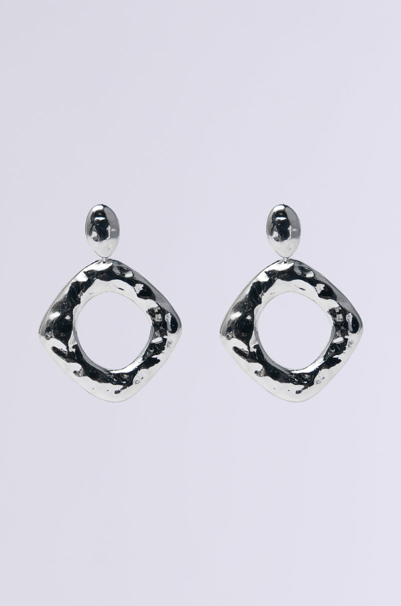 The NEVER BENT STATEMENT EARRING features faux silver geometric design with a hammered texture, creating a striking look. Each earring consists of a small circular stud connected to a larger hollow diamond shape against a plain white background, highlighting their elegance.