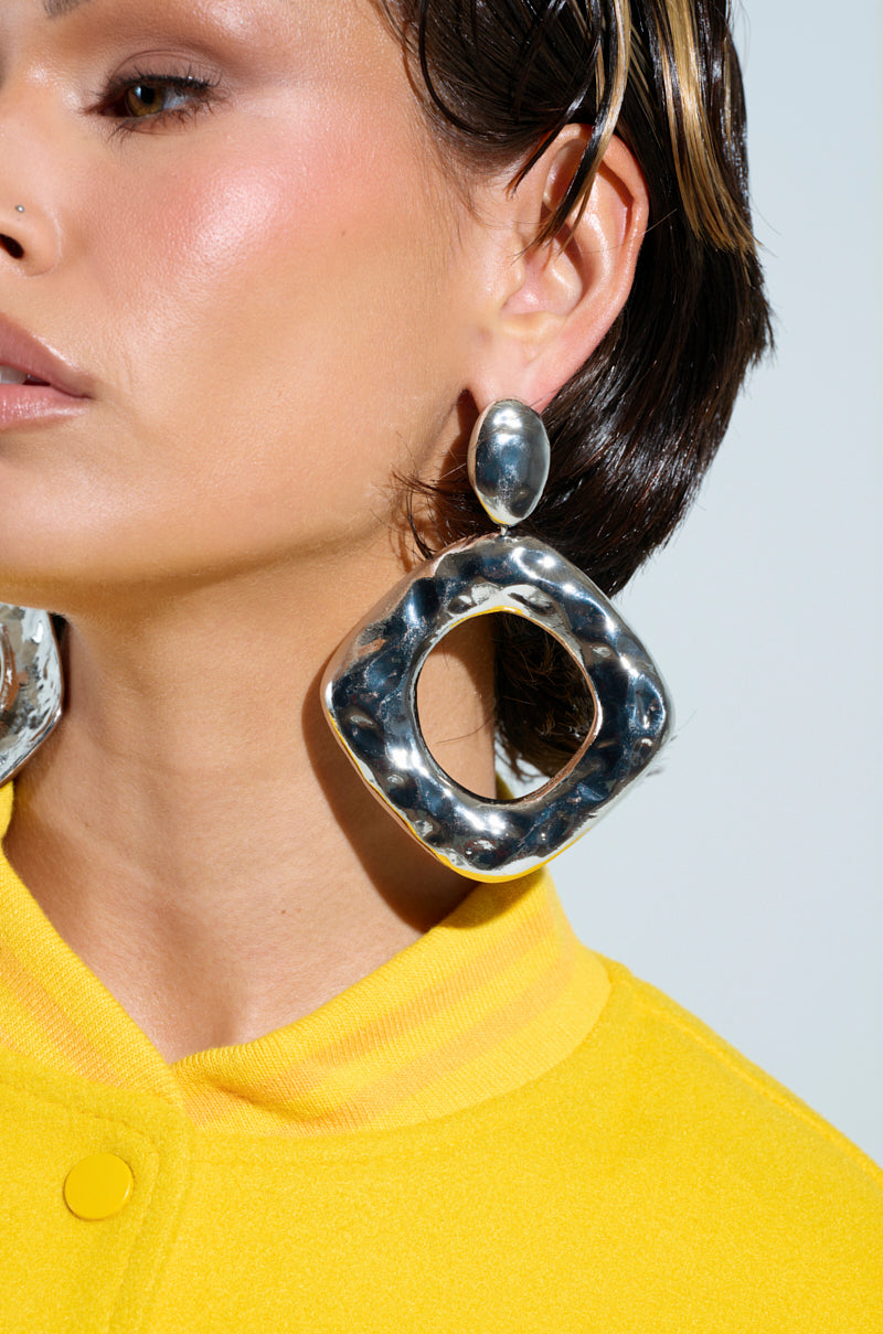 A close-up features someone wearing the NEVER BENT STATEMENT EARRING, a large textured square with rounded corners in faux silver. It's paired with a bright yellow top and styled-back hair against a plain light backdrop.