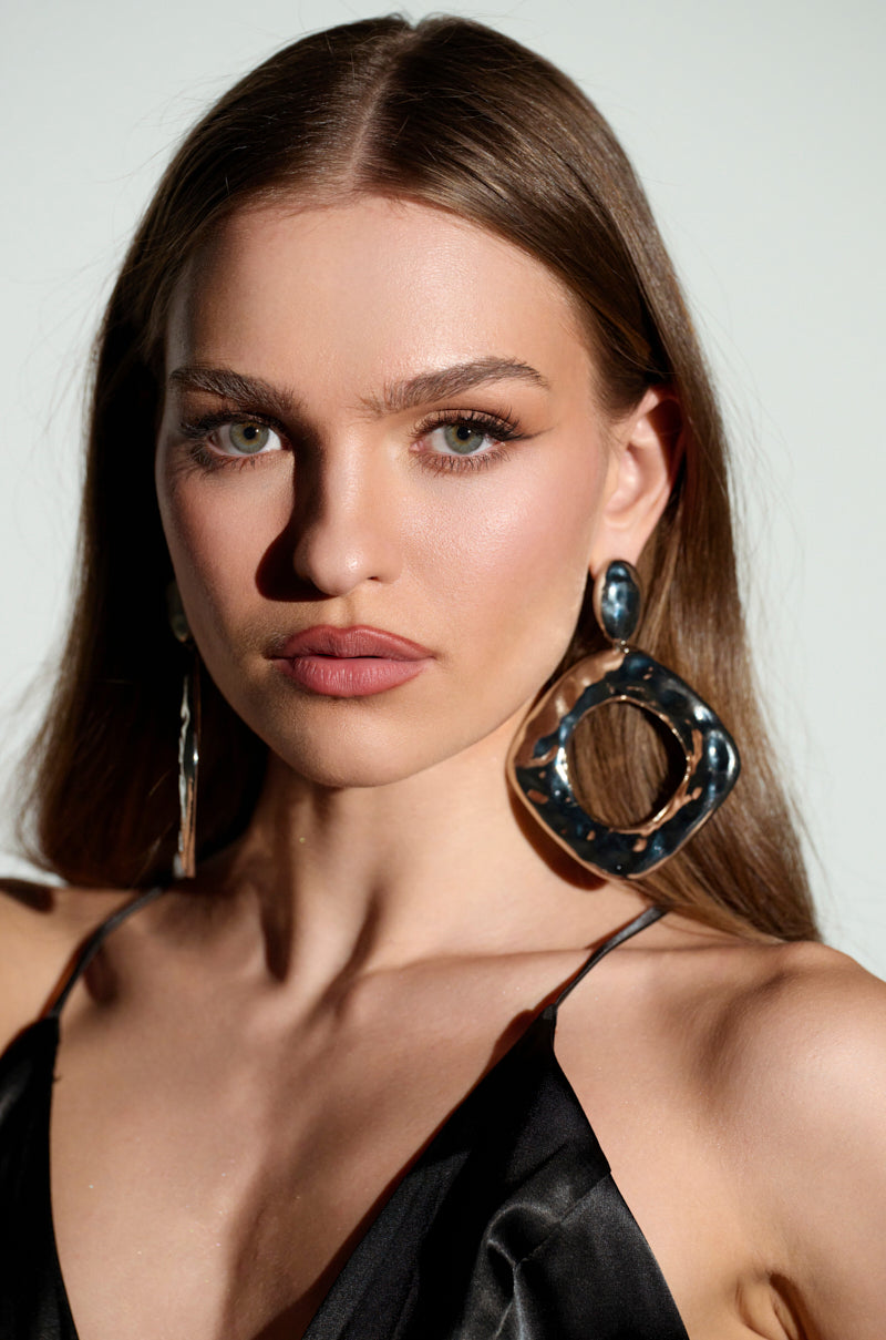 A woman with long brown hair in a black top poses against a light background. She confidently wears large NEVER BENT STATEMENT EARRINGS, while the sunlight casts gentle shadows on her face.