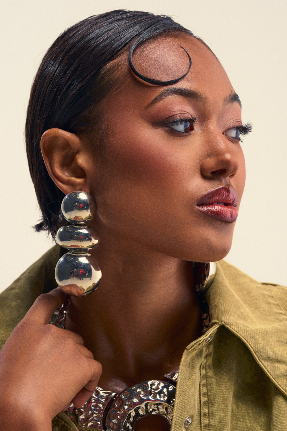A woman with slicked-back hair and bold eye makeup wears statement jewelry, including the BALLS TO THE WALL EARRING—large, reflective metallic silver earrings—and a gold jacket. She gazes thoughtfully to the side, her hand resting near her face.