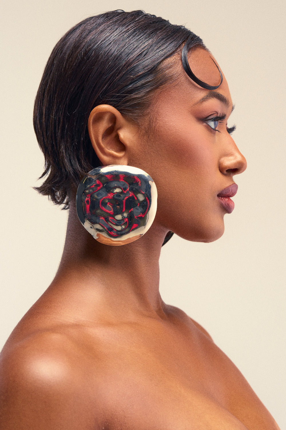 A woman with smooth, dark skin and short, sleek hair poses in profile, wearing the BEND MY WAY EARRING—an oversized disc earring in bold black, red, and cream hues that serves as a striking statement piece. Her makeup is natural against a neutral background.