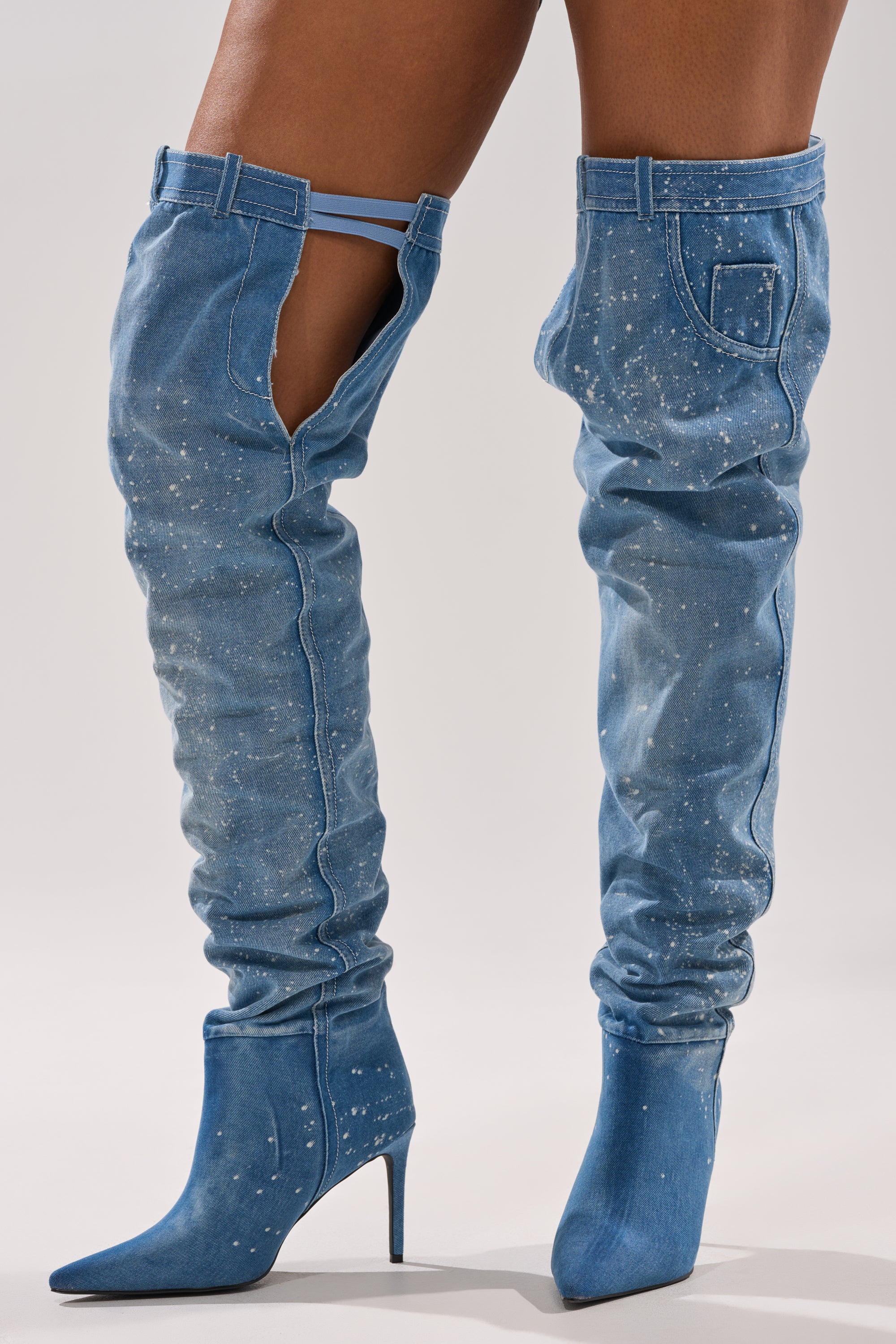 Close-up of a person wearing the AZALEA WANG JEANNI DENIM SPLATTER PAINTED STILETTO BOOT, featuring thigh-high length, white paint splatter, a pocket detail, and an open side cutout, against a plain gray background.