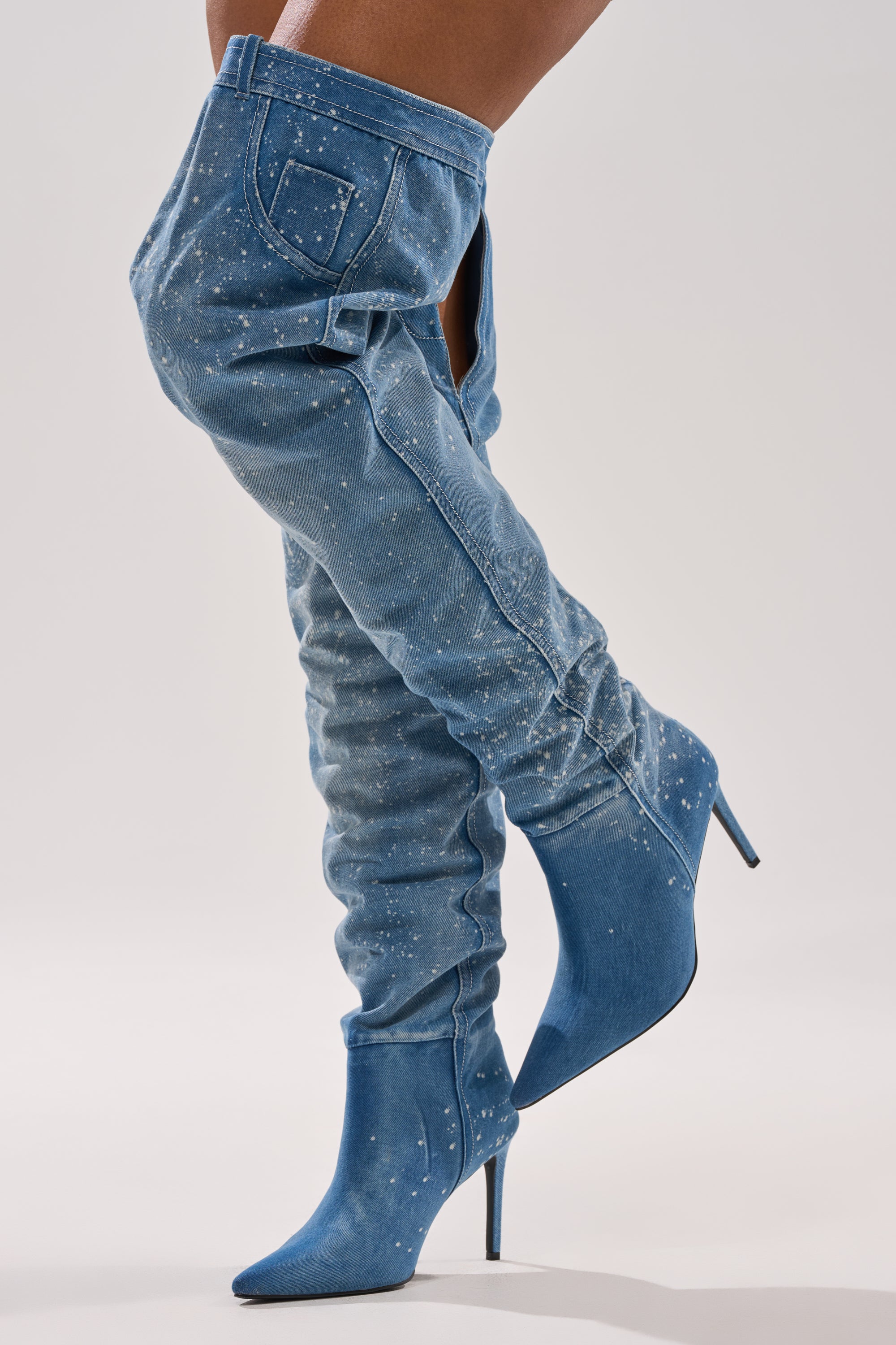 Someone wears the AZALEA WANG JEANNI DENIM SPLATTER PAINTED STILETTO BOOT, a thigh-high stiletto with pointed toes, jean-inspired pockets, and paint splatter details, shown against a plain, light-colored background.