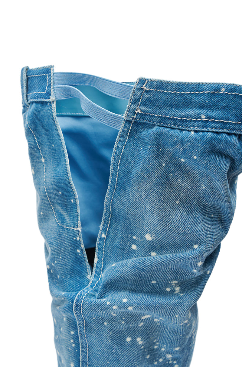 A close-up of the AZALEA WANG JEANNI DENIM SPLATTER PAINTED STILETTO BOOT showcases blue denim with paint splatter detailing and a partially unzipped design revealing a light blue inner waistband and fabric.