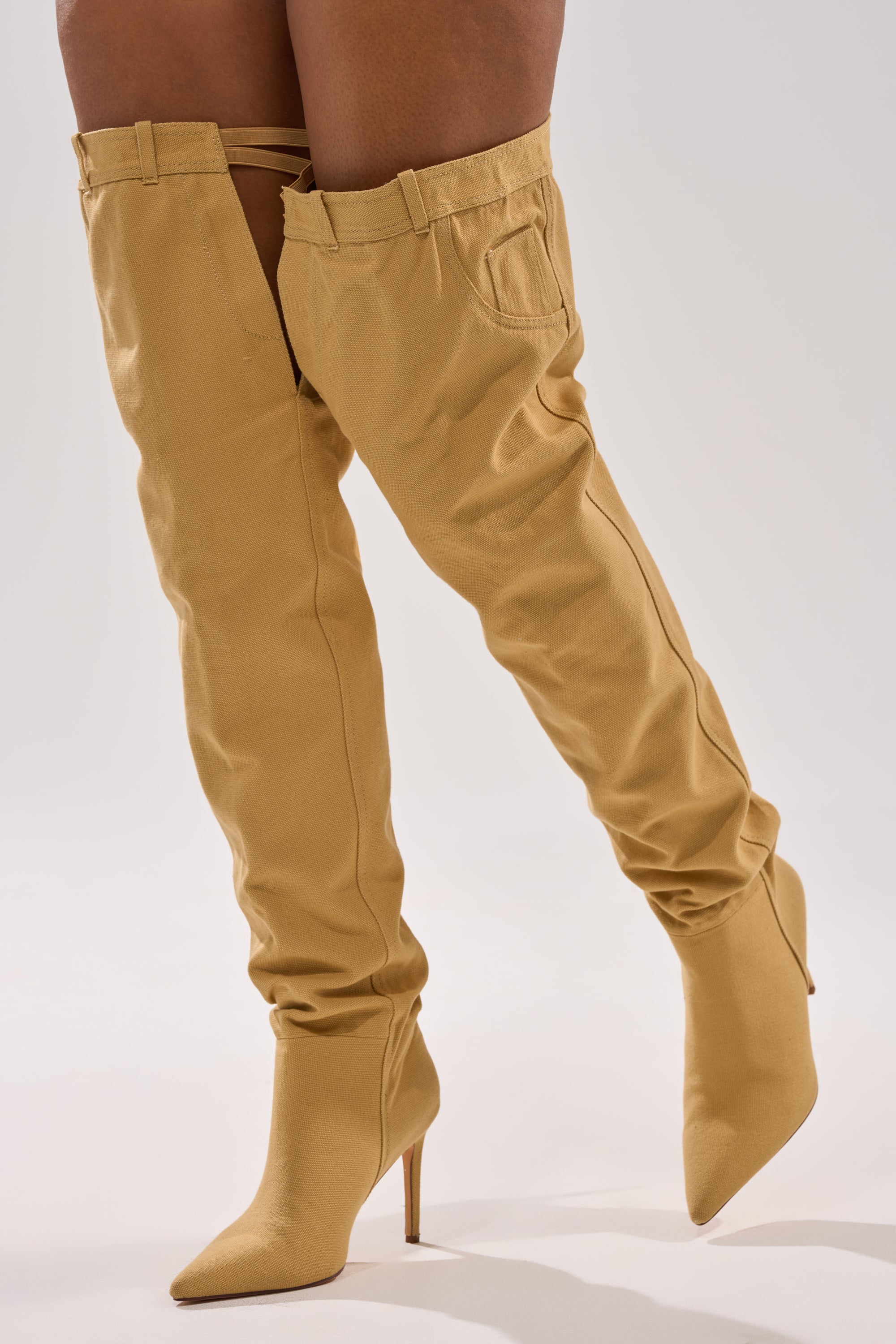 A person wearing the AZALEA WANG JEANNI TAN PANT DETAIL OVER THE KNEE BOOT, which features stiletto heels, belt loops, and a back pocket to resemble pants, stands on a light background.