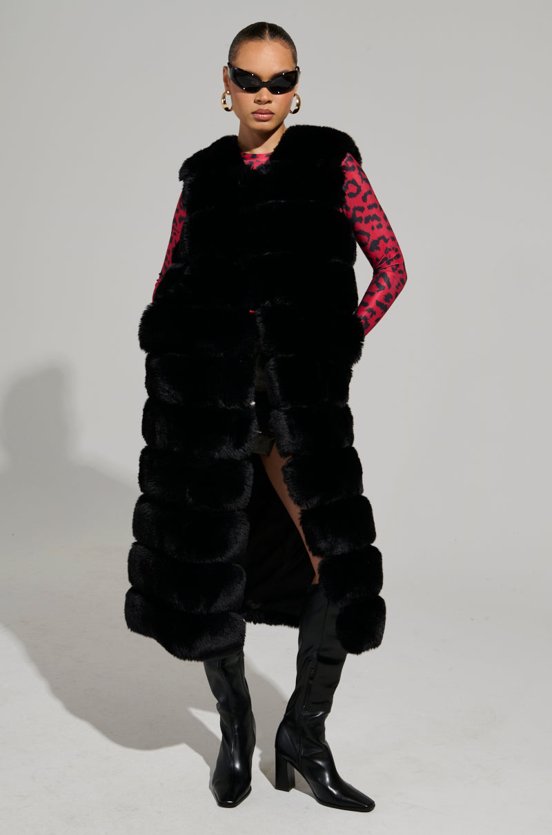 A person wearing the JENNY LIN FAUX FUR MAXI VEST IN BLACK, paired with a red and black patterned top, black shorts, knee-high black boots, and sunglasses, stands confidently against a plain background. This ensemble is the epitome of a chic winter wardrobe.