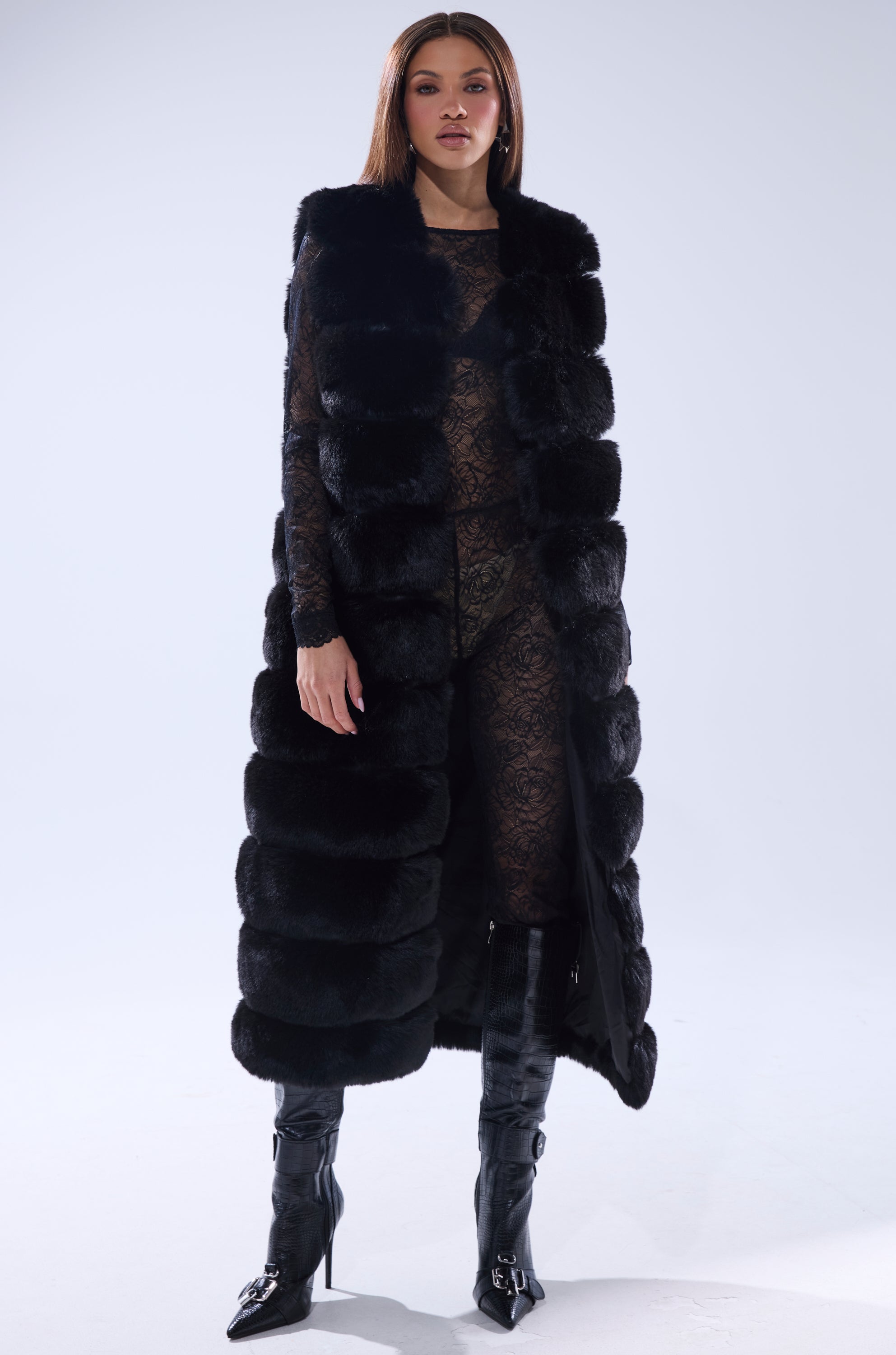 A woman faces forward in the JENNY LIN FAUX FUR MAXI VEST IN BLACK, styled over a sheer lacy outfit with knee-high boots, posing against a plain light background—an elegant showcase of winter outerwear.