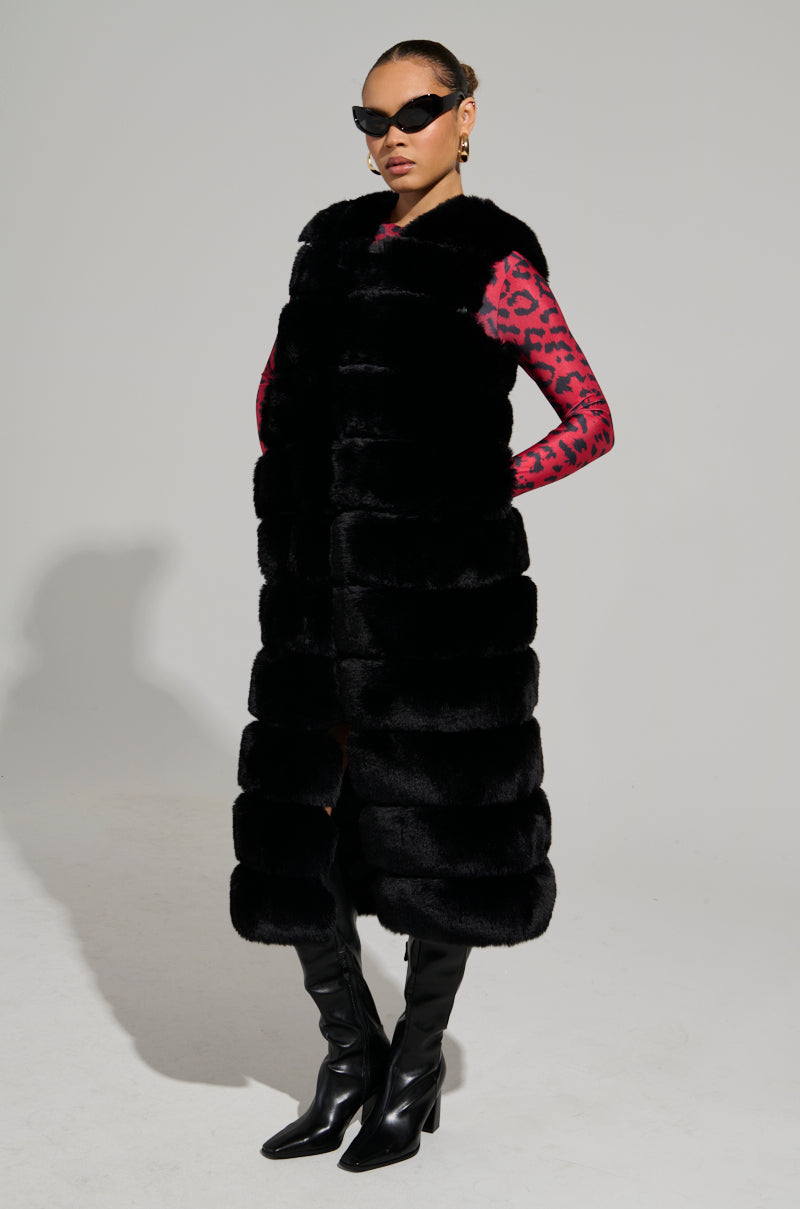 Sporting a pair of sunglasses and the JENNY LIN FAUX FUR MAXI VEST IN BLACK over a pink and black patterned top, the individual confidently poses in tall black boots against a plain background—an essential look for any stylish winter wardrobe.