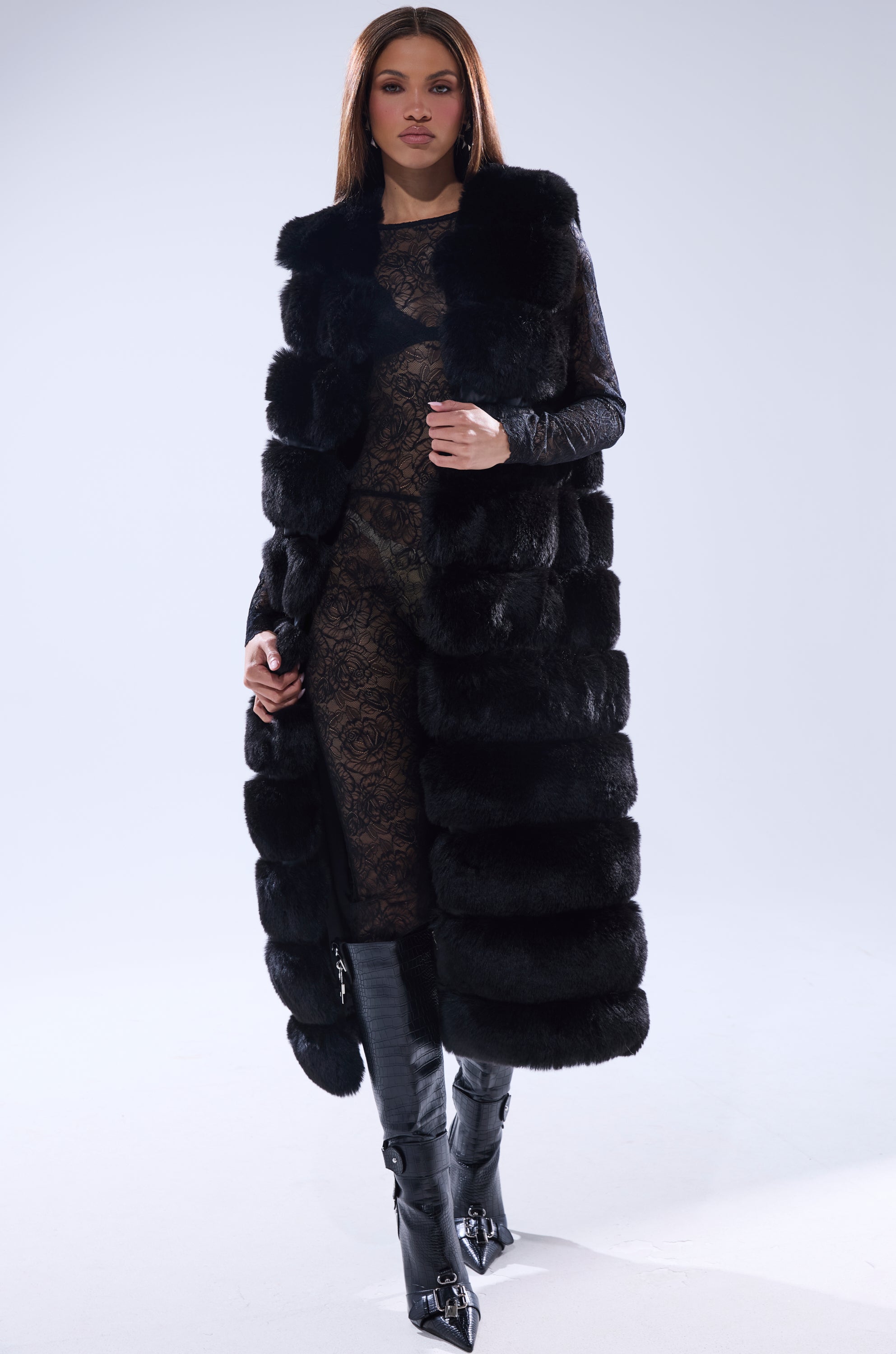 A woman stands confidently against a plain light background, showcasing the JENNY LIN FAUX FUR MAXI VEST IN BLACK layered over a sheer lace dress and paired with sleek black knee-high boots.