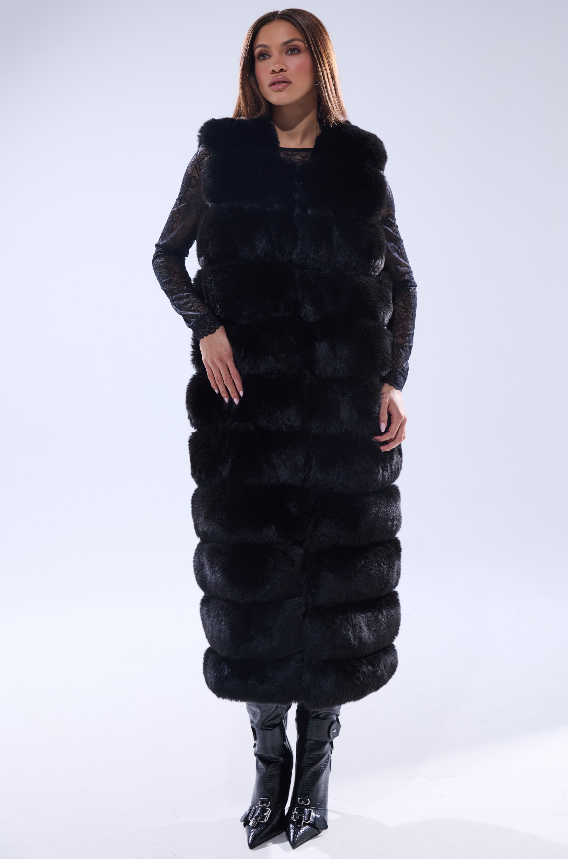 A woman with straight brown hair and a neutral expression models the JENNY LIN FAUX FUR MAXI VEST IN BLACK over a black lace top and pointed-toe boots against a light background, showcasing chic winter outerwear.