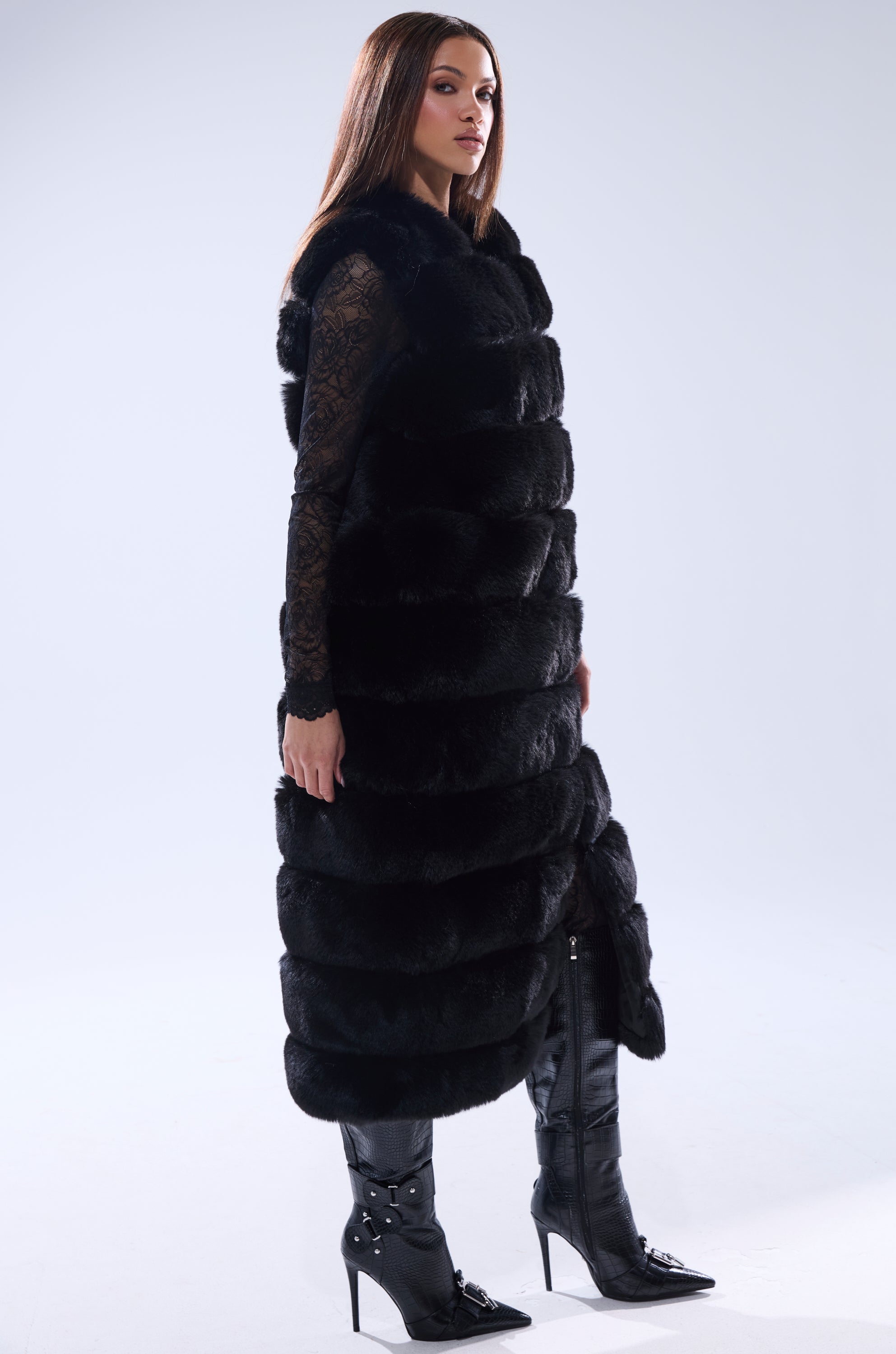 A woman wearing the JENNY LIN FAUX FUR MAXI VEST IN BLACK over a lace top and knee-high heeled boots stands against a plain light background, looking confidently to the side.