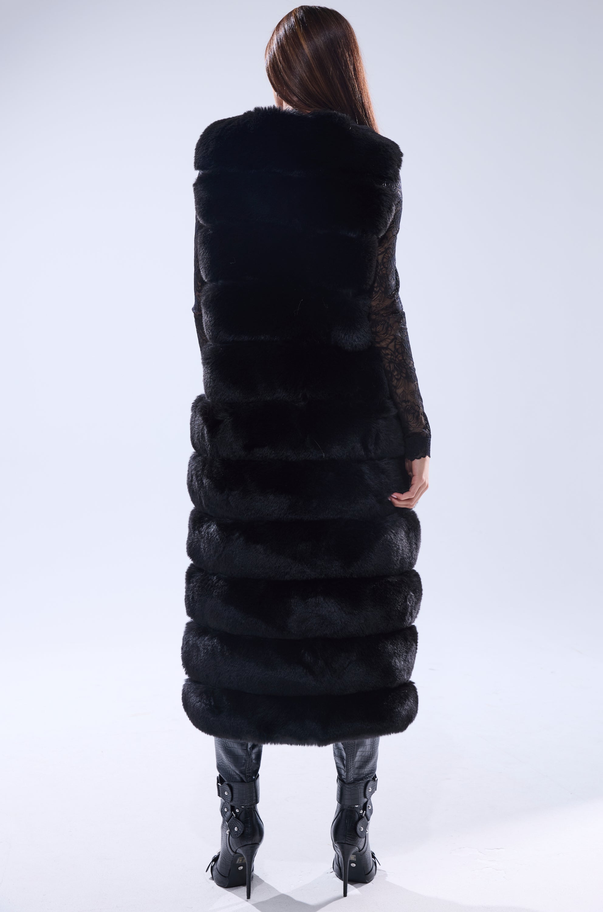 A person with long, straight brown hair faces away from the camera, modeling the JENNY LIN FAUX FUR MAXI VEST IN BLACK and high-heeled boots against a simple light background.