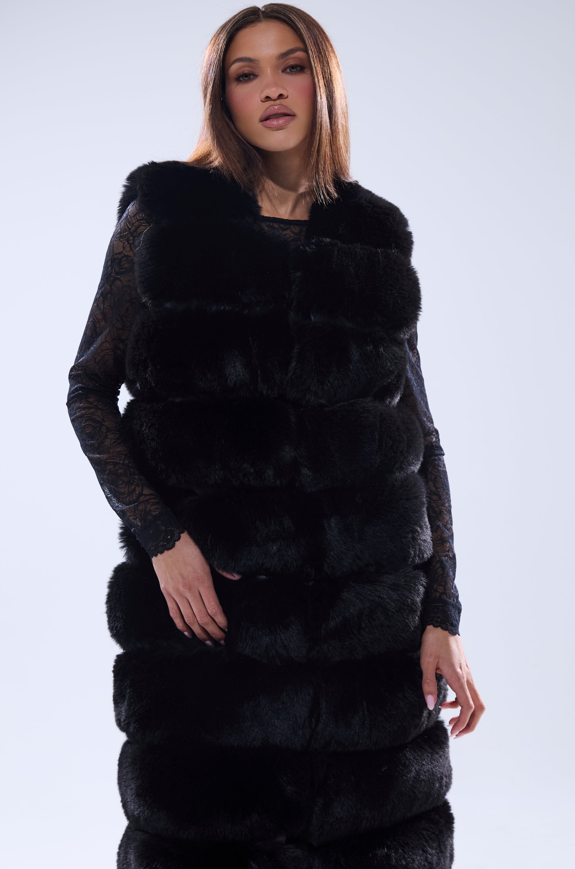 Wearing the JENNY LIN FAUX FUR MAXI VEST IN BLACK over a sheer lace top, a person with straight brown hair poses against a light background, delivering an effortlessly chic winter look while gazing at the camera.
