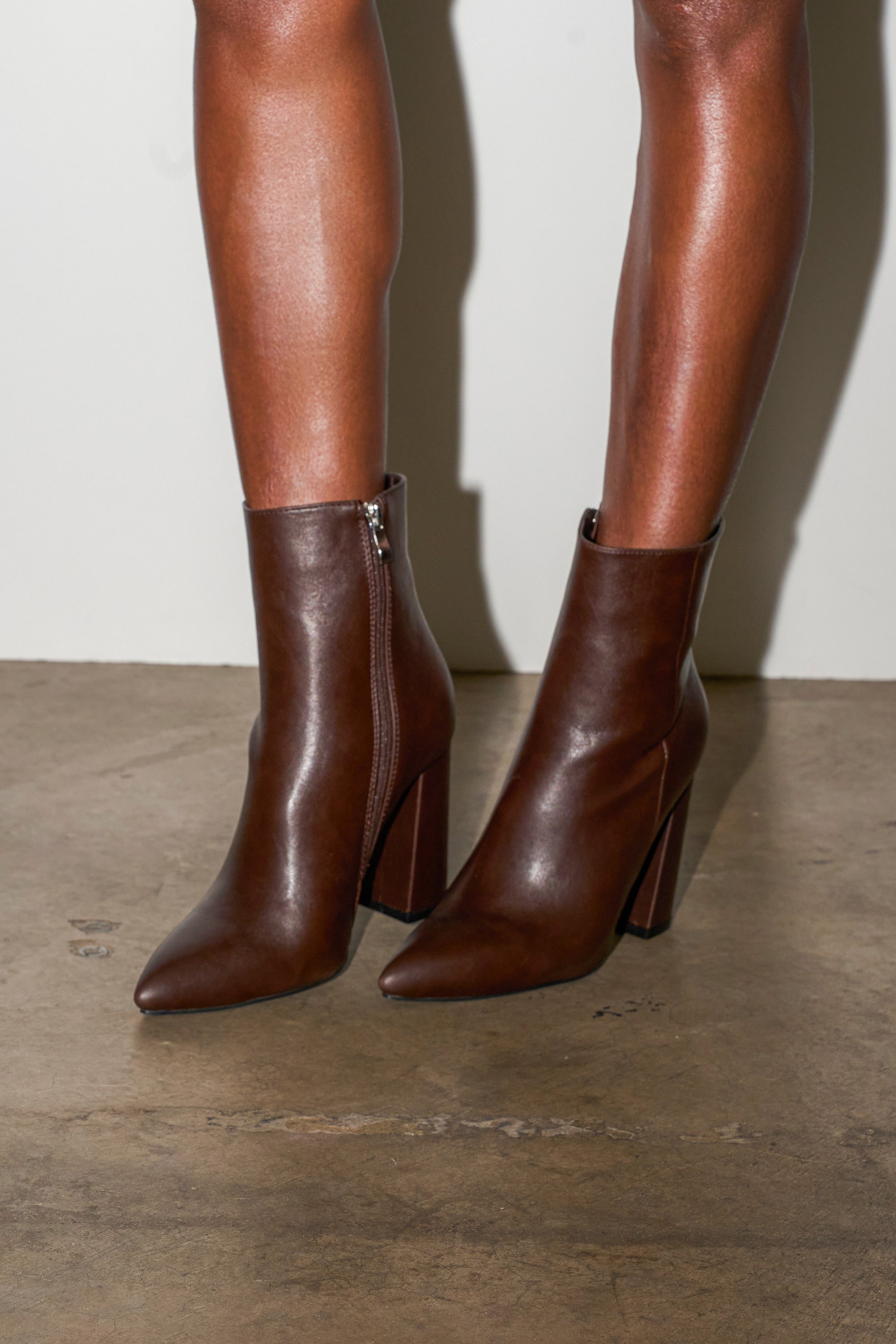 Someone stands on a concrete floor against a plain white wall, wearing the AZALEA WANG JESSABELL Brown Pointed Toe Bootie with side zippers.