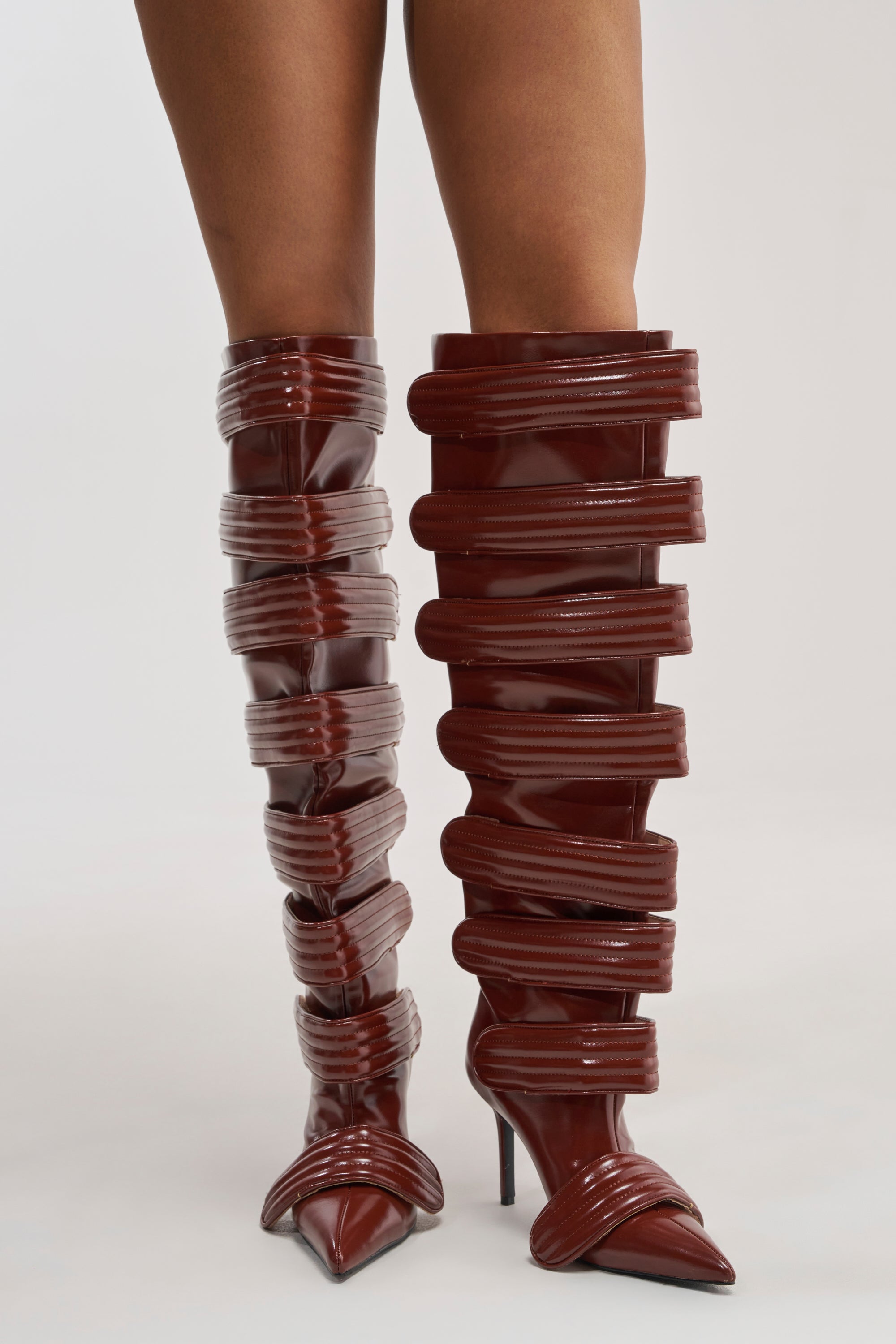 A person wearing the AZALEA WANG JETSET COGNAC STILETTO BOOT, featuring glossy, dark red faux leather with multiple horizontal padded bands and pointed toes, stands against a plain white background.