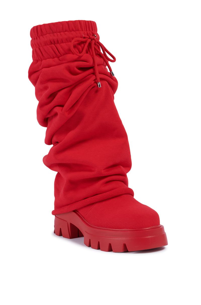 The AZALEA WANG JETSETTER RED BOOT features a slouchy, scrunched-up design with a chunky platform lug sole for enhanced traction. This boot includes a stretchy, sweatpant-inspired drawstring closure at the top and is crafted from soft, flexible material, offering a casual and contemporary look.