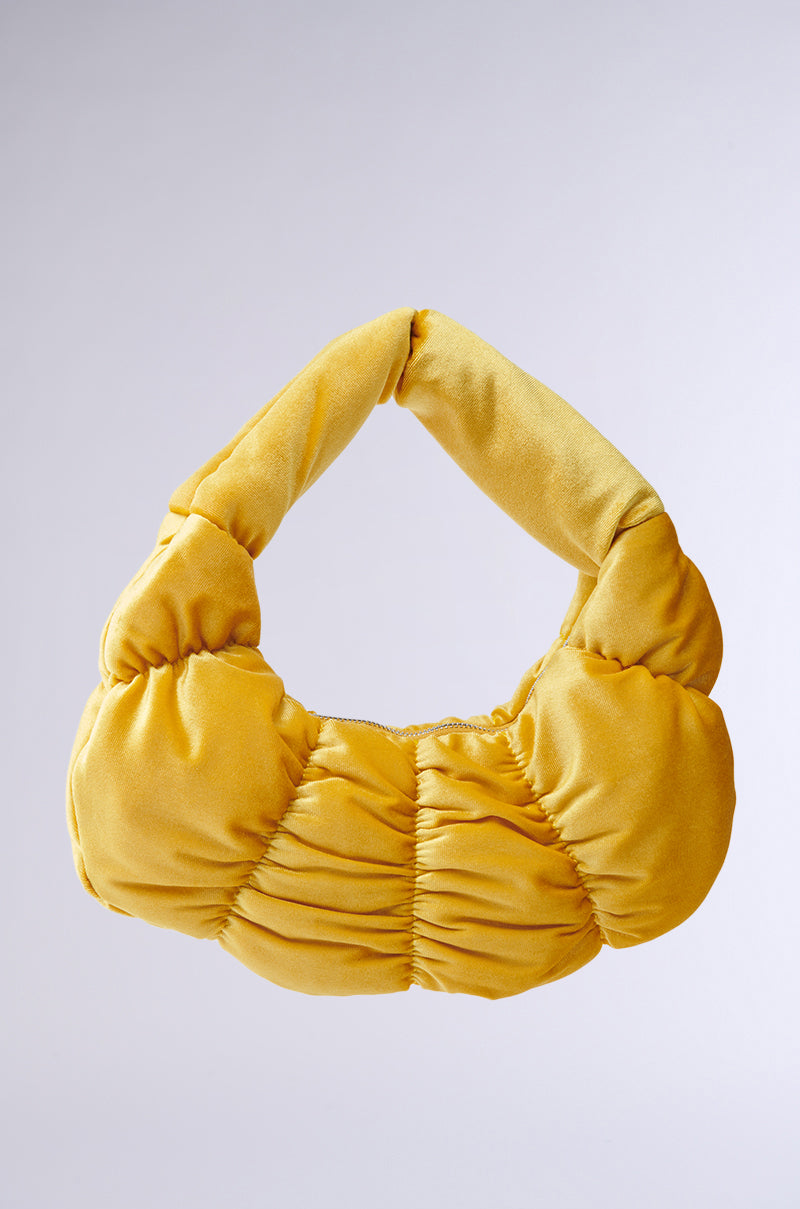 The TRENDY ONE VELVET RUCHED PURSE is a yellow, crescent-shaped handbag with a soft quilted texture and single handle. It features a metallic zip-top closure and is highlighted against a plain gray background. 1