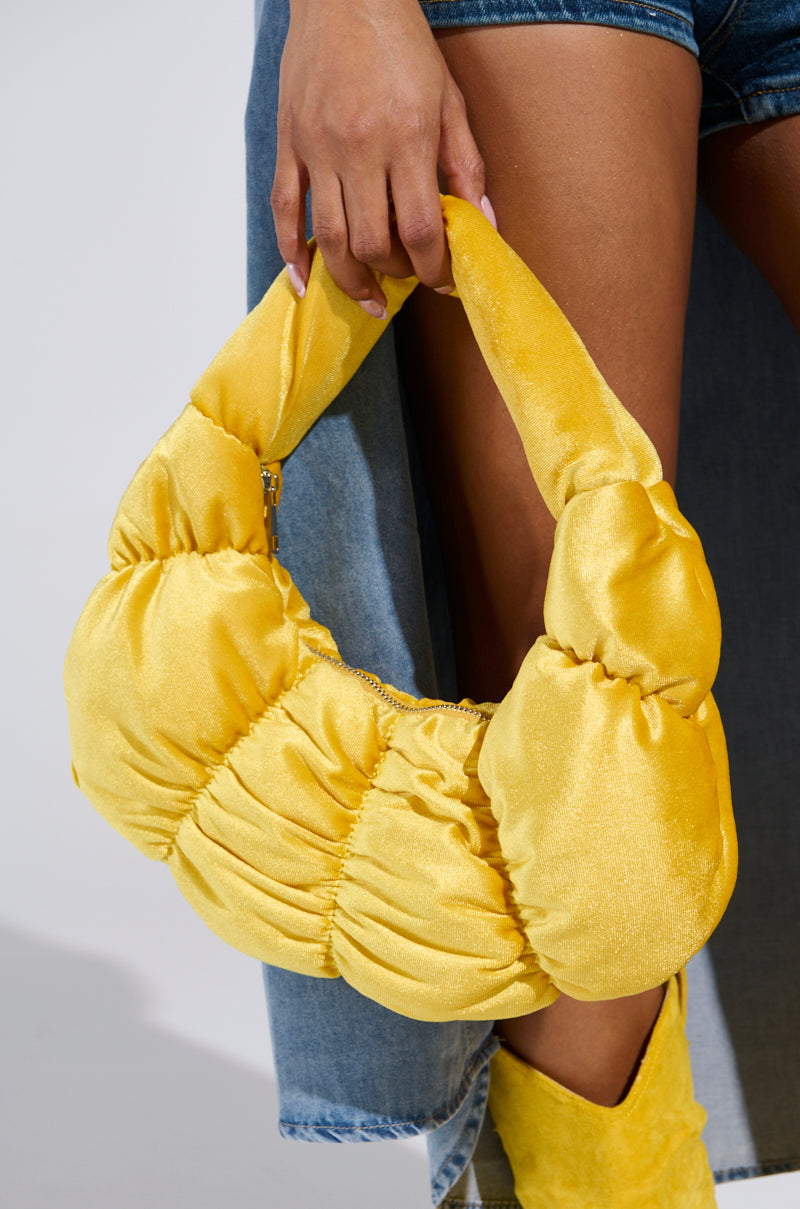 Dressed in denim shorts and a long denim jacket, a person carries the TRENDY ONE VELVET RUCHED PURSE, a puffy yellow satin handbag with a metallic zip-top closure. Yellow suede boots complete the look in bright lighting that enhances the vibrant textures and colors. 2