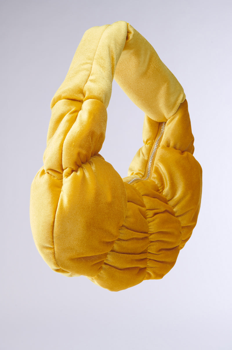 A mustard yellow, plush headband featuring a twisted crescent shape with a side zipper is set against a plain gray backdrop. 3