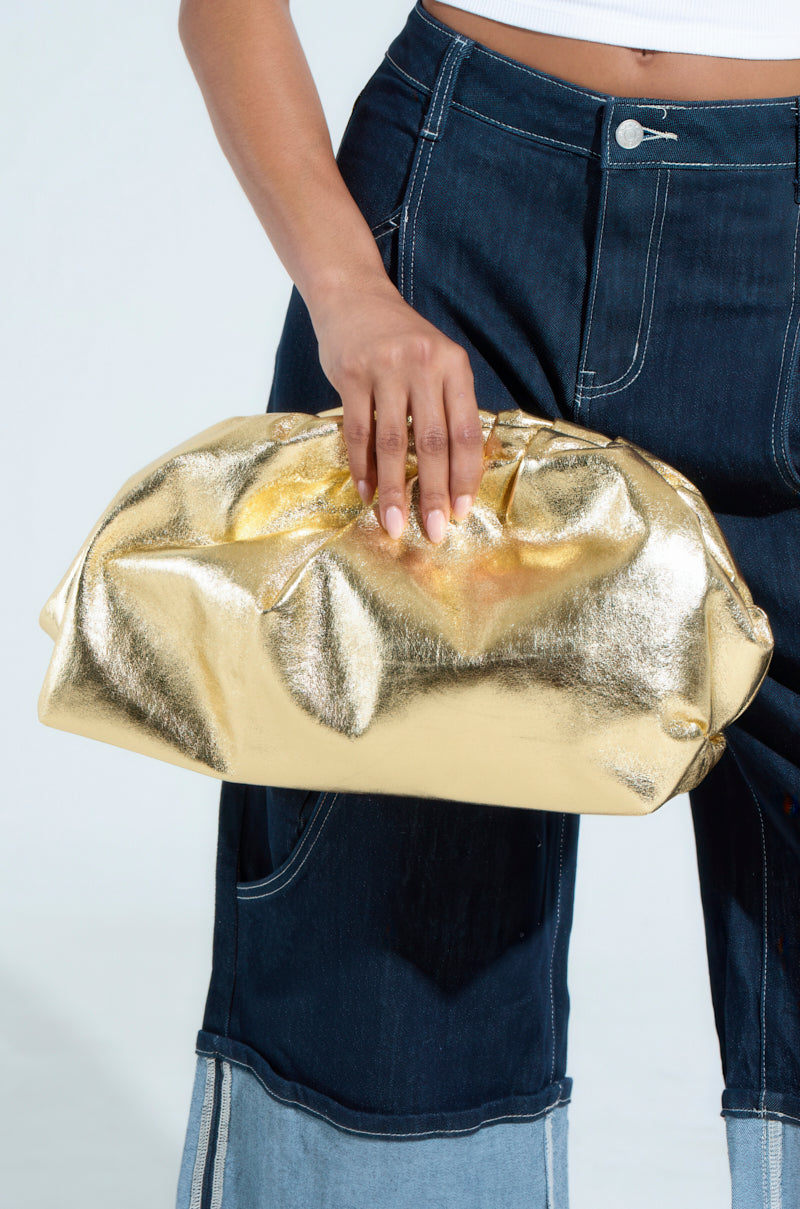 A person holds the OVER IT METALLIC CLUTCH against dark denim pants with white stitching, a glimpse of a crisp white top showing, all set against a plain light background. 1