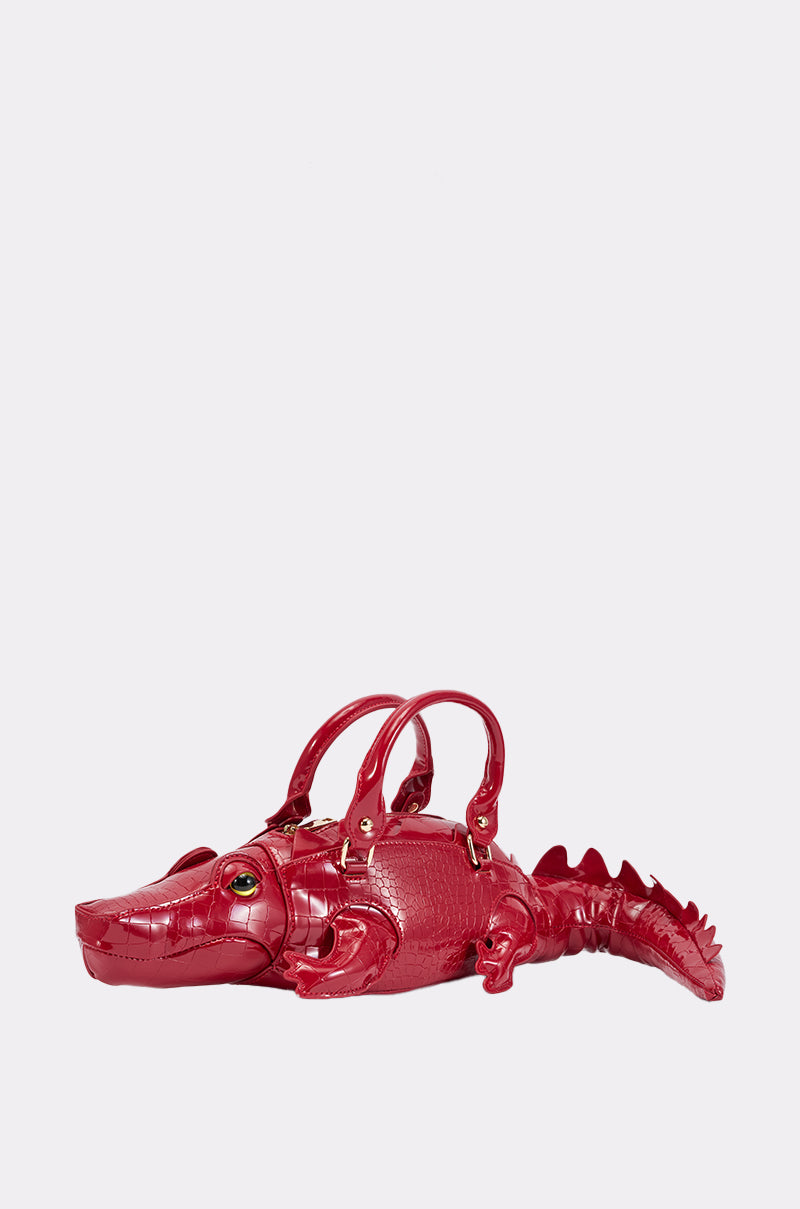 The ALLIGATOR BAG is a shiny red croc-embossed faux leather purse featuring gold hardware and two handles, photographed against a plain light background.