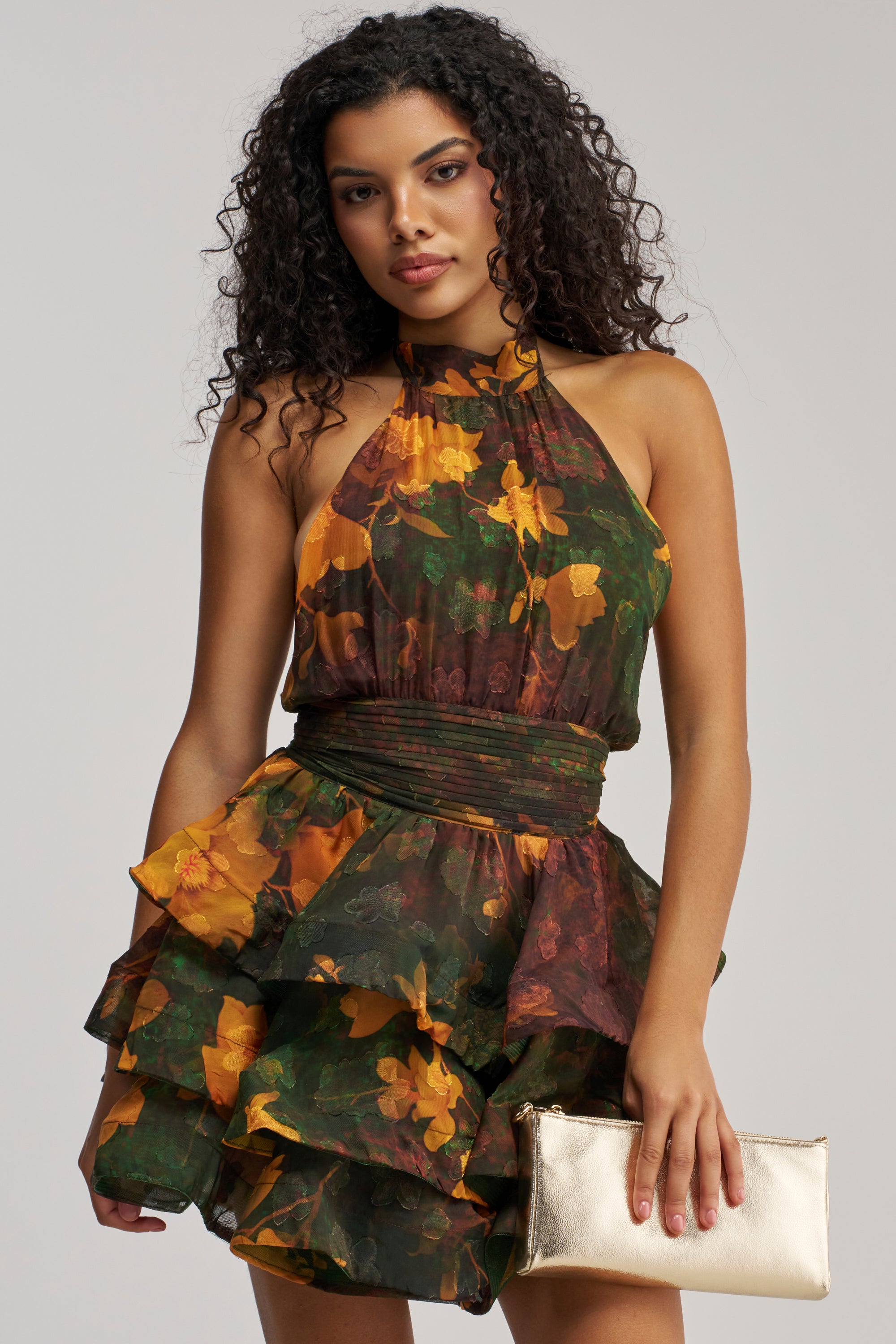 A woman with curly hair wears the SPIN ME AROUND CHIFFON MINI DRESS IN GREEN FLORALS — a sleeveless, ruffled mini dress featuring a dark green and yellow floral print. She holds a metallic gold clutch bag and stands against a plain light background. This dress is perfect for summer date nights or as an elegant wedding guest outfit.