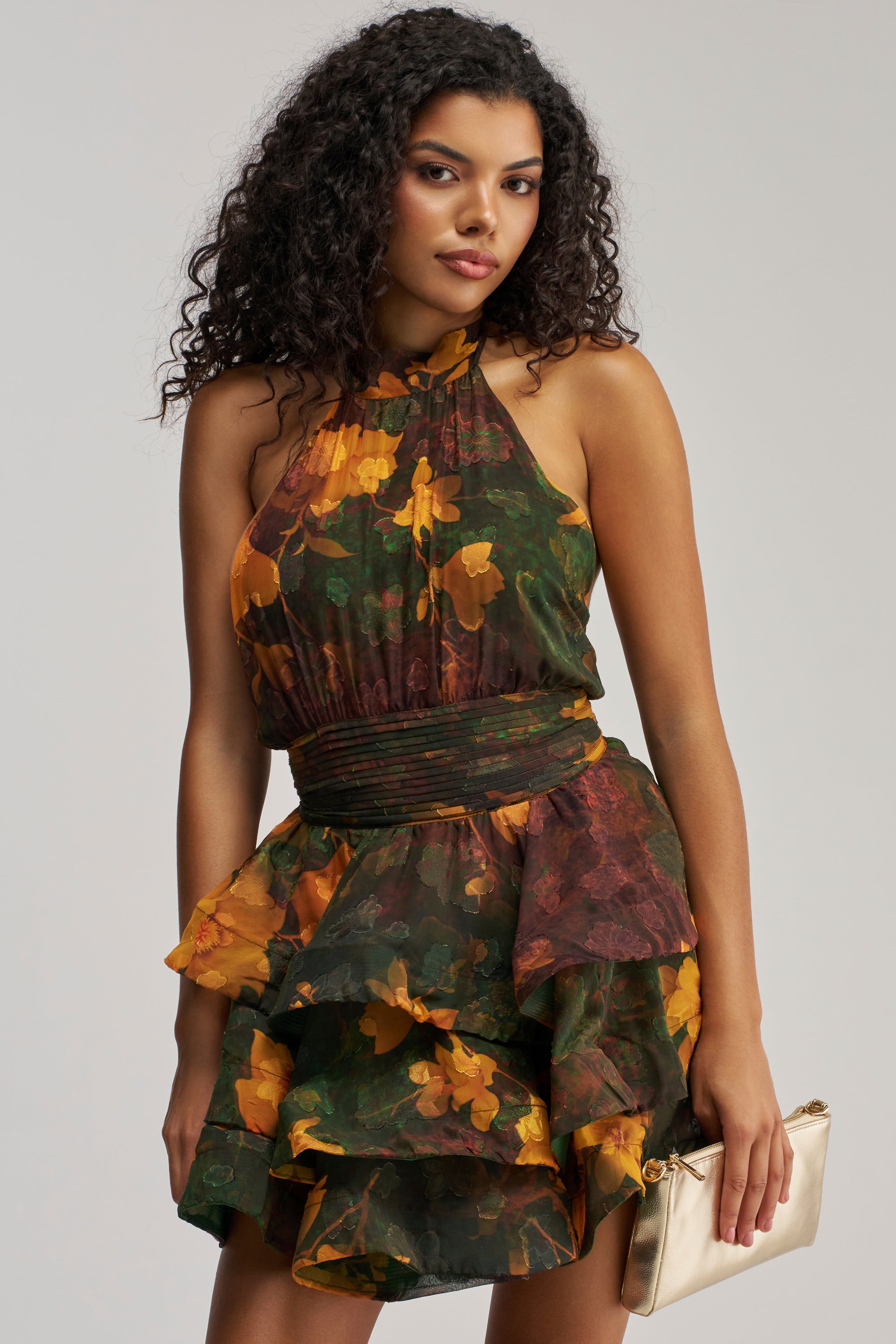 Wearing the SPIN ME AROUND CHIFFON MINI DRESS IN GREEN FLORALS—an elegant sleeveless mini dress featuring ruffled details and a vibrant dark green and yellow floral chiffon design—a woman with curly dark hair holds a metallic gold clutch against a plain background, creating a chic look perfect for summer date nights or as a stylish wedding guest dress.