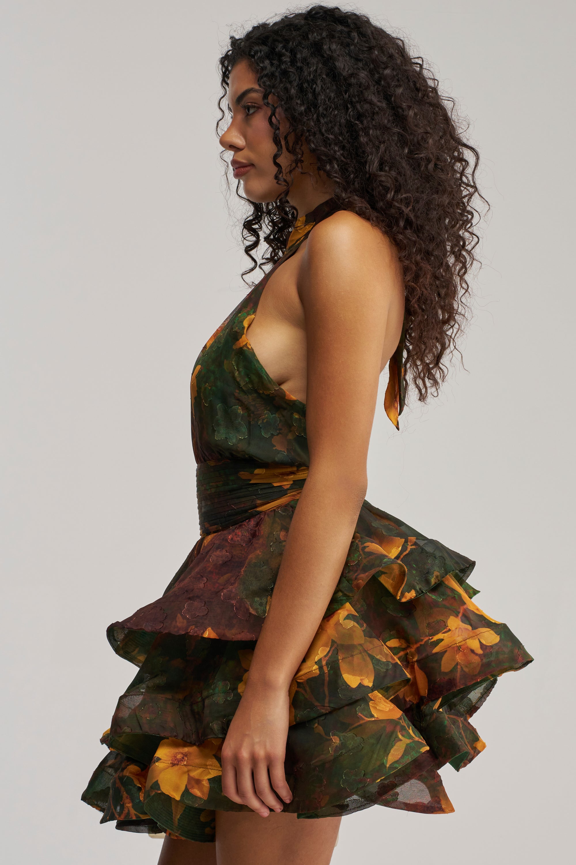 A woman with curly hair stands in profile against a plain light background, wearing the SPIN ME AROUND CHIFFON MINI DRESS IN GREEN FLORALS—a sleeveless mini dress featuring green and brown floral chiffon, yellow floral accents, and a layered ruffled skirt—perfect for summer date nights or as a chic wedding guest dress.
