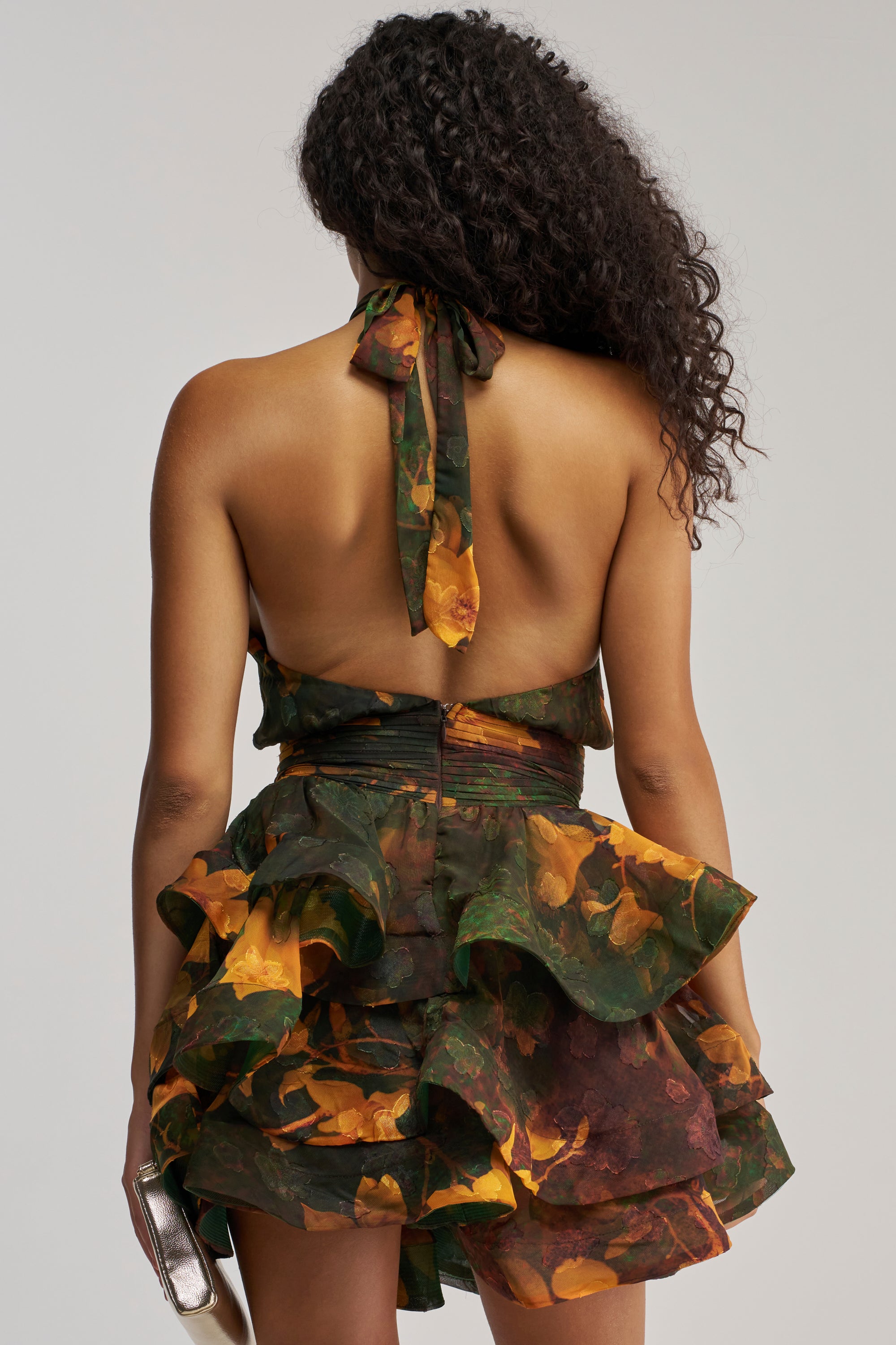 A woman with curly hair is seen from behind against a plain background, wearing the SPIN ME AROUND CHIFFON MINI DRESS IN GREEN FLORALS—a halter-style, backless mini dress featuring layered ruffles and a delicate floral chiffon fabric—while holding a small metallic clutch, creating an effortlessly chic look ideal for wedding guests or summer date nights.