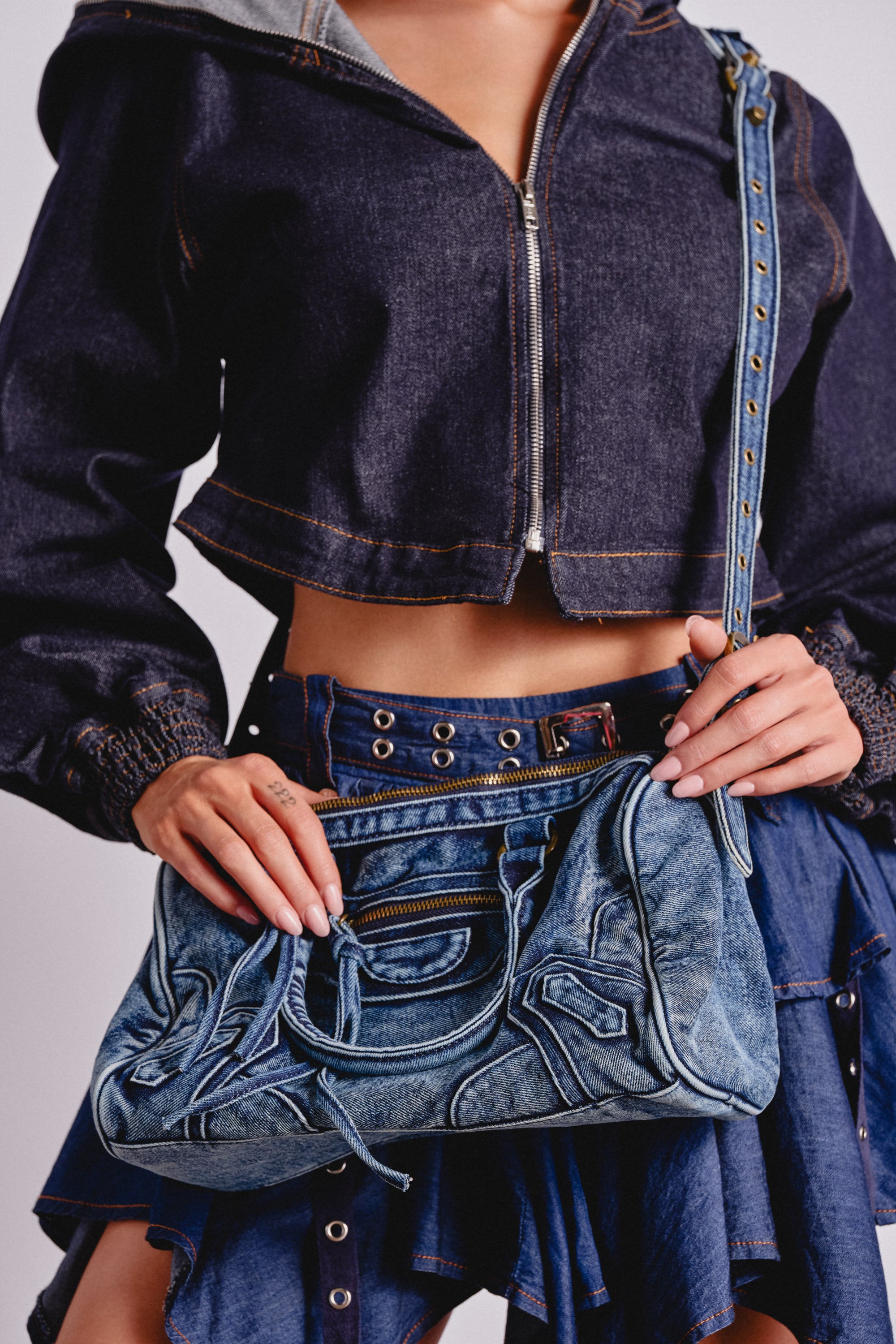 A woman holds the IM THAT GIRL DISTRESSED PURSE—a washed denim handbag—while wearing a cropped dark denim jacket and ruffled skirt. Only her torso and hands show against a plain background, highlighting this cool streetwear accessory. 0