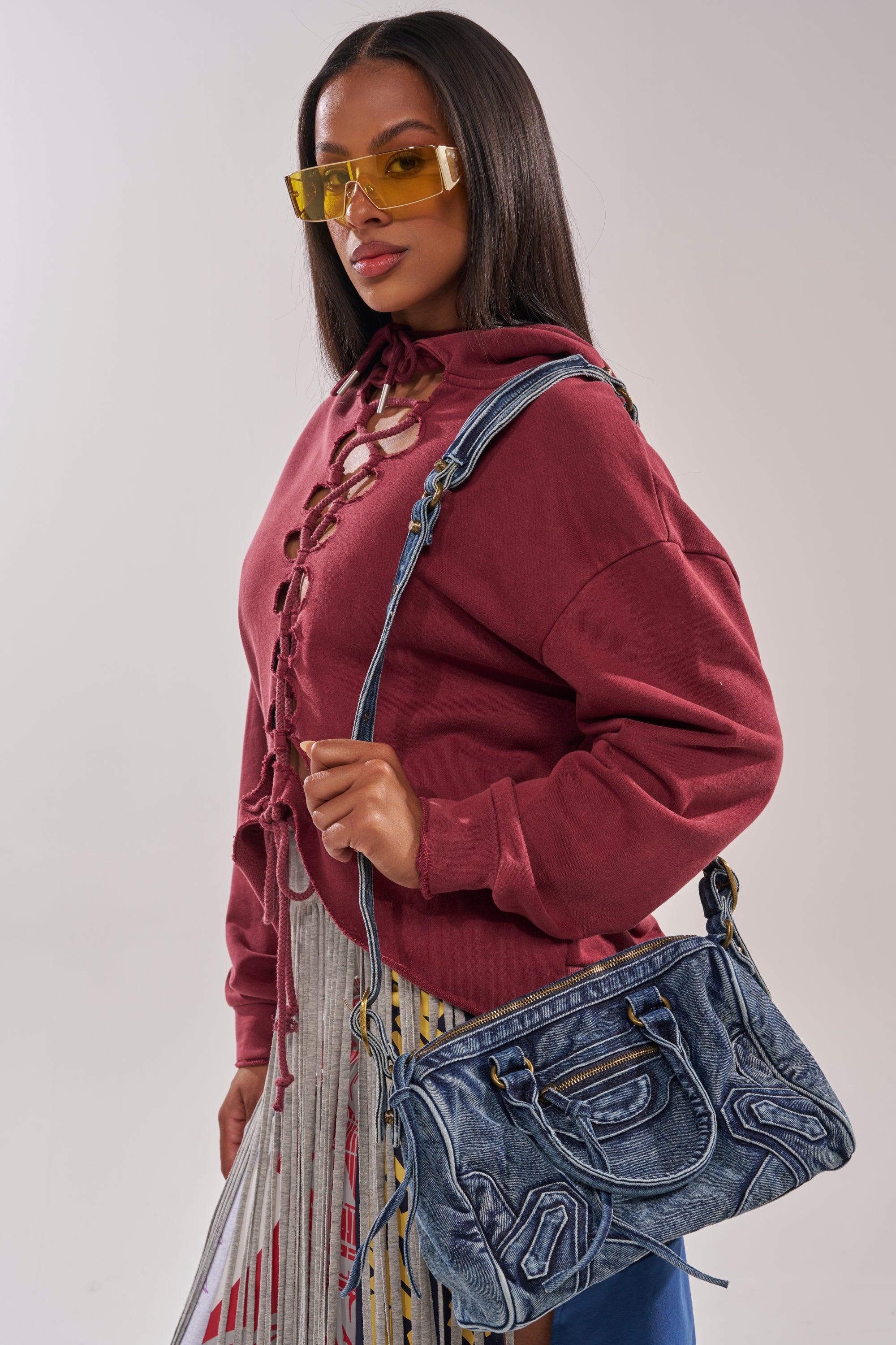A woman in yellow-tinted sunglasses and a burgundy lace-up top holds the IM THAT GIRL DISTRESSED PURSE, a washed denim handbag, confidently over her shoulder against a plain backdrop. 2