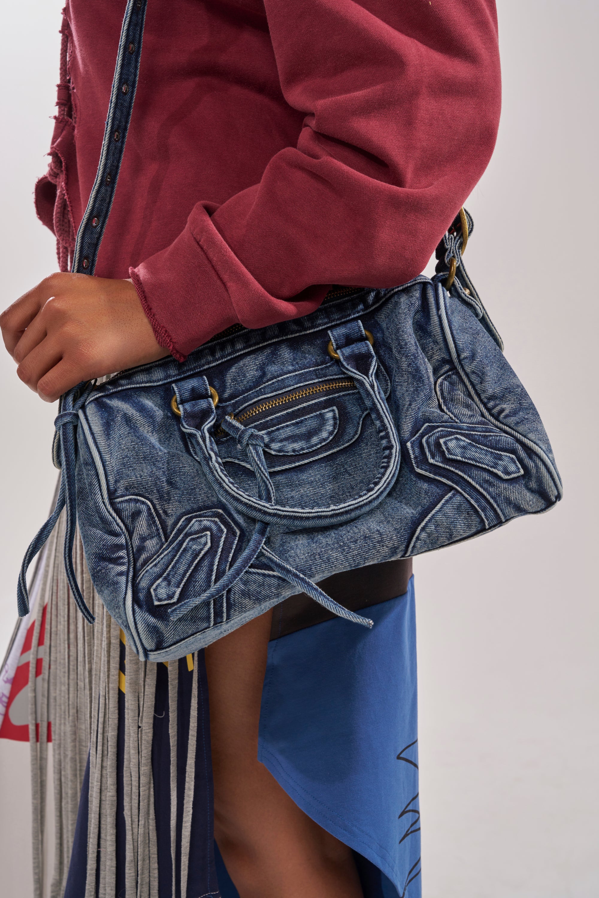 A person in a red long-sleeve top and color-blocked skirt carries the IM THAT GIRL DISTRESSED PURSE, a washed denim handbag with decorative stitching, zippered pocket, and matching strap for effortless style. 3