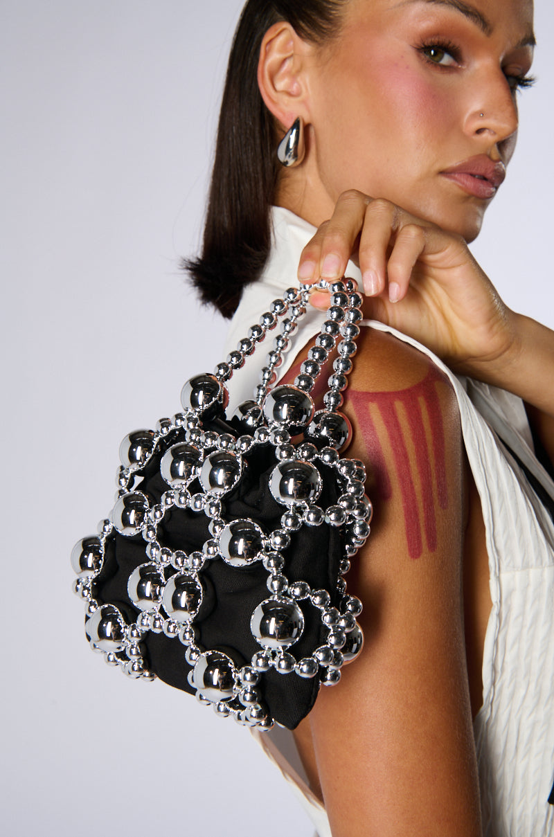 A woman holds the chic KEEP ME CLOSE CHROME PURSE, a black mini accessory decorated with a striking geometric pattern of metallic silver beads. She is dressed in a classy white outfit complemented by silver jewelry, and has three red vertical stripes painted on her shoulder. The scene is set against a plain light gray background.