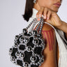 A woman holds the chic KEEP ME CLOSE CHROME PURSE, a black mini accessory decorated with a striking geometric pattern of metallic silver beads. She is dressed in a classy white outfit complemented by silver jewelry, and has three red vertical stripes painted on her shoulder. The scene is set against a plain light gray background.