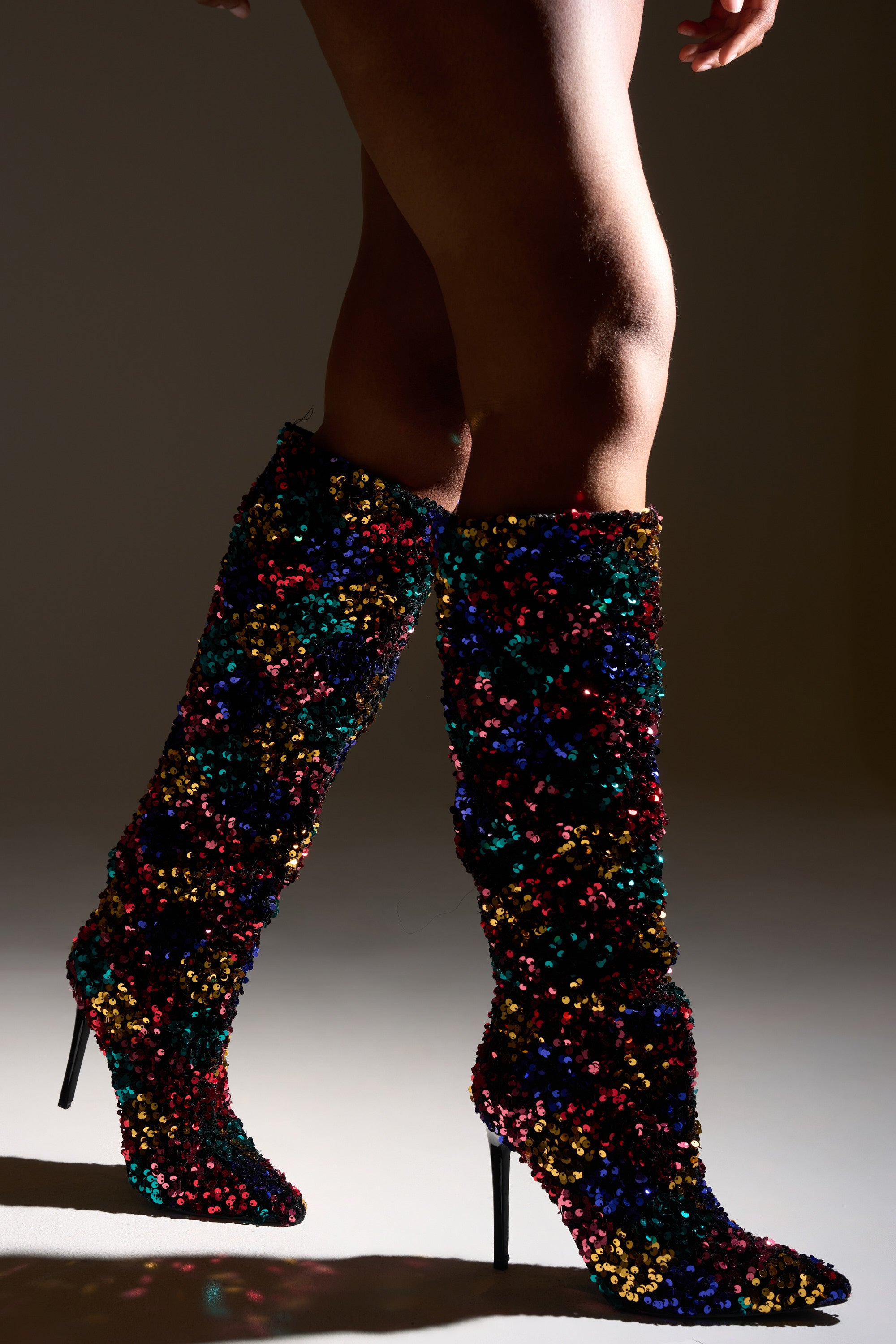 A person models the AZALEA WANG JIMENA MULTI SEQUIN EMBELLISHED BOOT—knee-high, pointed-toe stiletto boots in vibrant multicolor sequins—shining against a softly lit backdrop with only their legs and the dazzling boots visible. 2